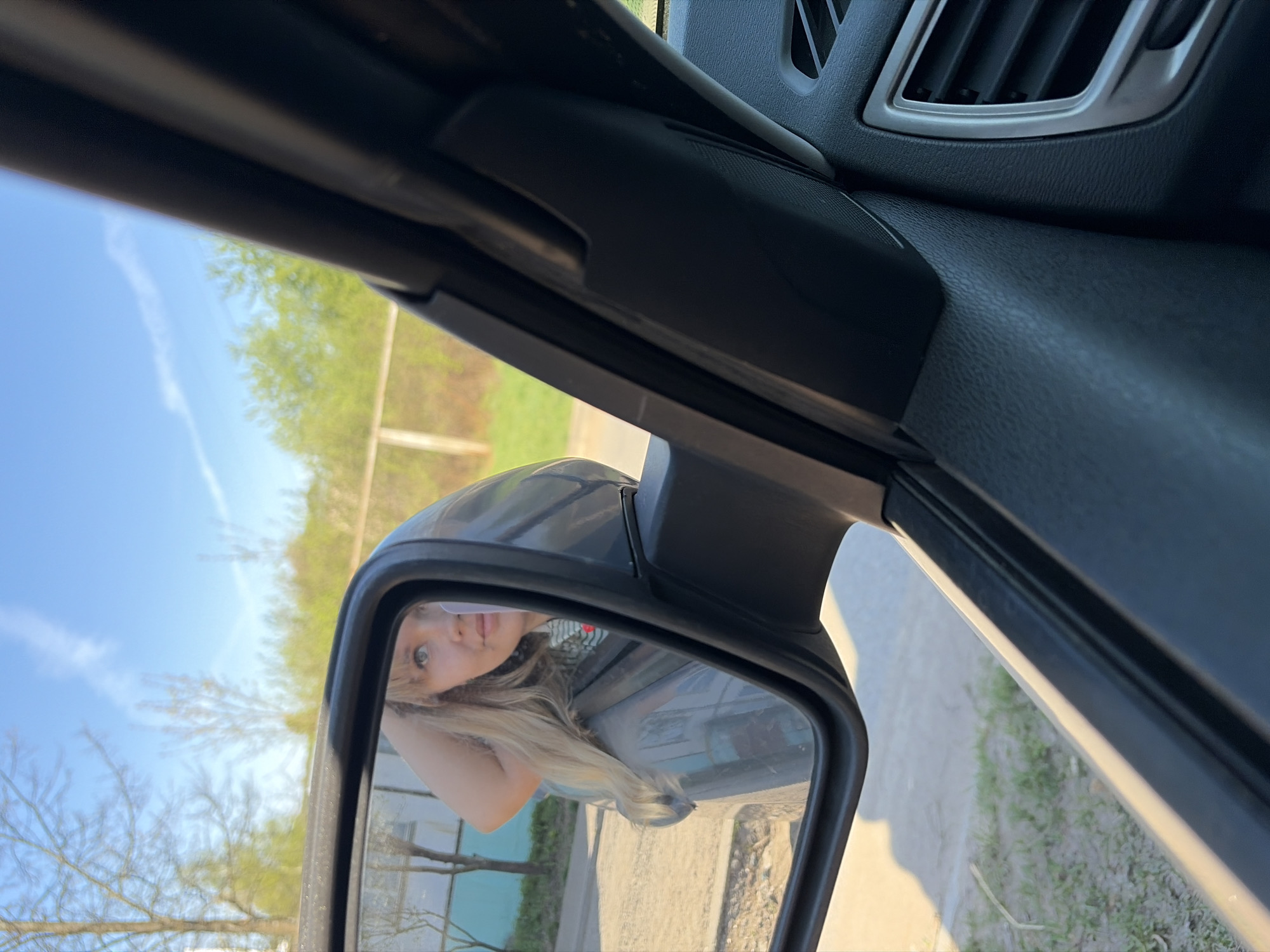 The image shows the interior of a car, looking towards the driver's side mirror. The mirror reflects a woman with blonde hair, and the external environment, including trees, sky, and a street. The car's interior components are visible, including the air vent and the door frame. It's impossible to determine the location to be Chaikovsky, Russia, based on the information provided. The scene appears to be outdoors, likely during the day, with the woman in the car looking towards the mirror.