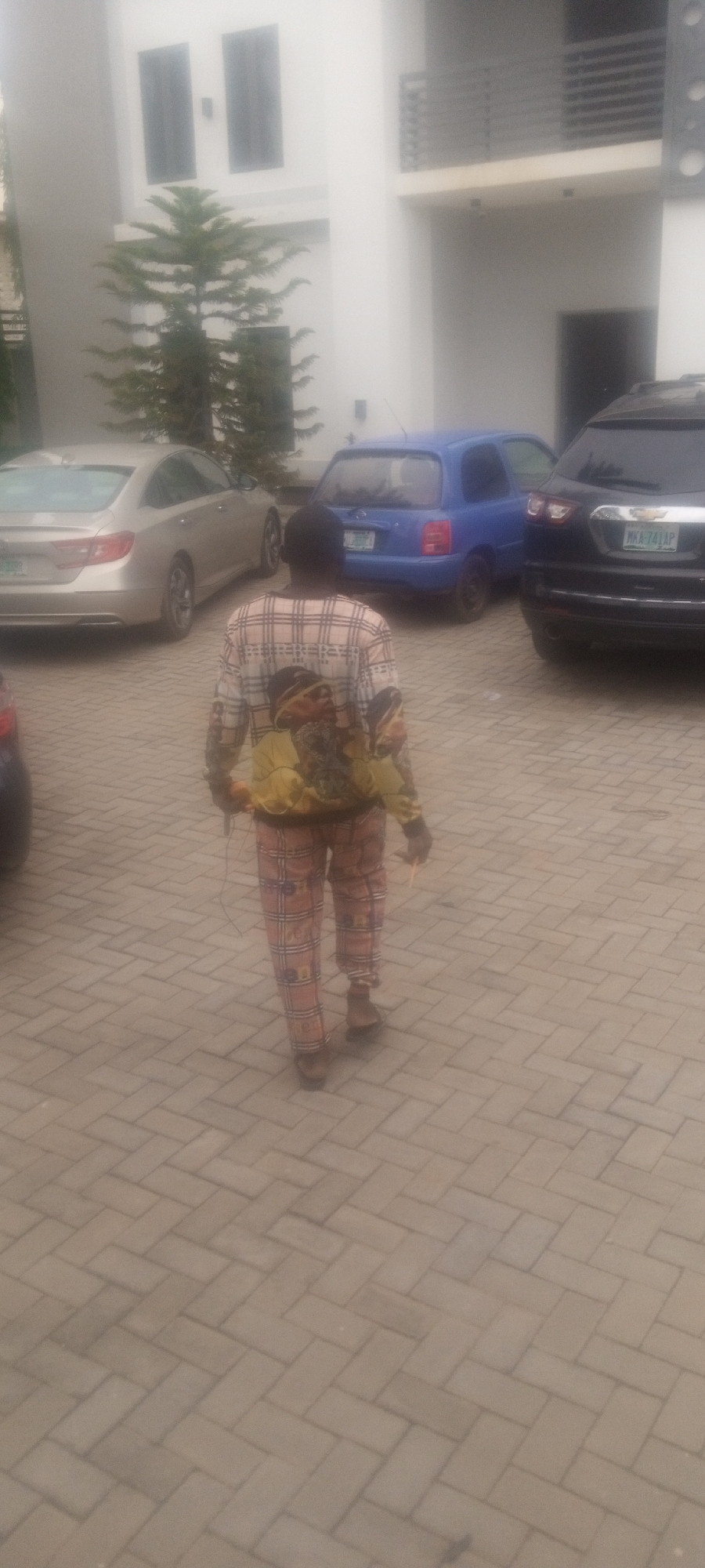 A person is walking away from the camera on a paved surface. The person is wearing a matching long-sleeved shirt and pants with a repeating checkered pattern and a depiction of two faces on the upper back. Behind the person are three cars parked in front of a two-story white building, including a beige sedan on the left, a small blue car in the center, and a dark-colored SUV on the right. There is a coniferous tree next to the building. The license plate on the SUV reads "MKA-747AP". The location is Unguwar Rimi, Nigeria.