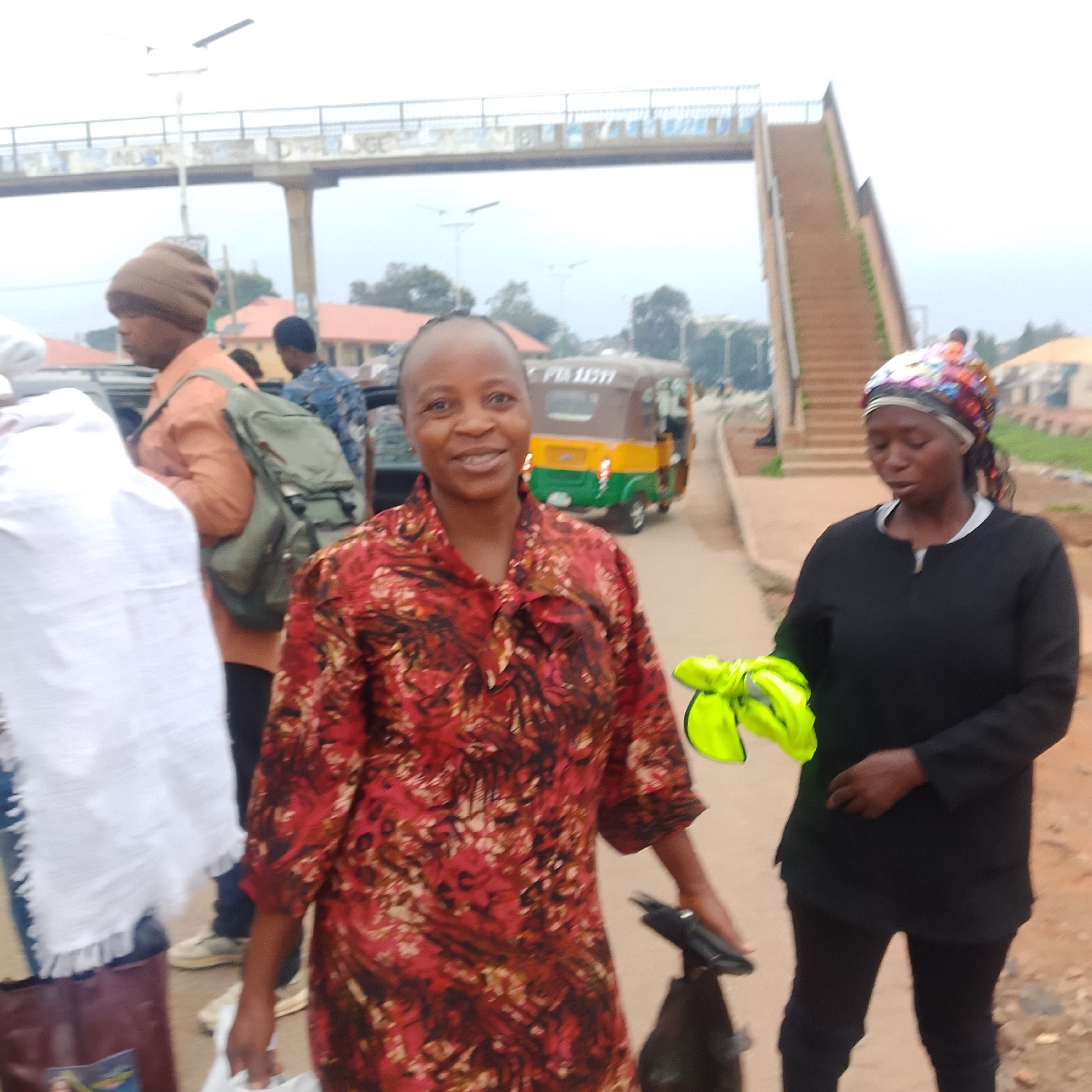 In the city of Unknown, Unknown, three individuals are pictured outdoors. The foreground is occupied by two women. The woman on the left, who is closer to the camera, is smiling and wearing a red and brown floral print shirt. She is holding a white bag in her left hand and a black object, possibly a spray bottle, in her right. To her right stands another woman wearing a black top and dark pants. She is holding a bright yellow vest in her hands. In the background, a man wearing an orange shirt and a beanie is visible, with a green backpack on. Behind him, another person in a blue patterned shirt is partially visible. An auto-rickshaw, yellow and green, is parked on the right side of the road, with a sign that reads "PTA 11377". An elevated pedestrian bridge is seen in the background, spanning across the scene. The ground appears to be a dirt or gravel road.