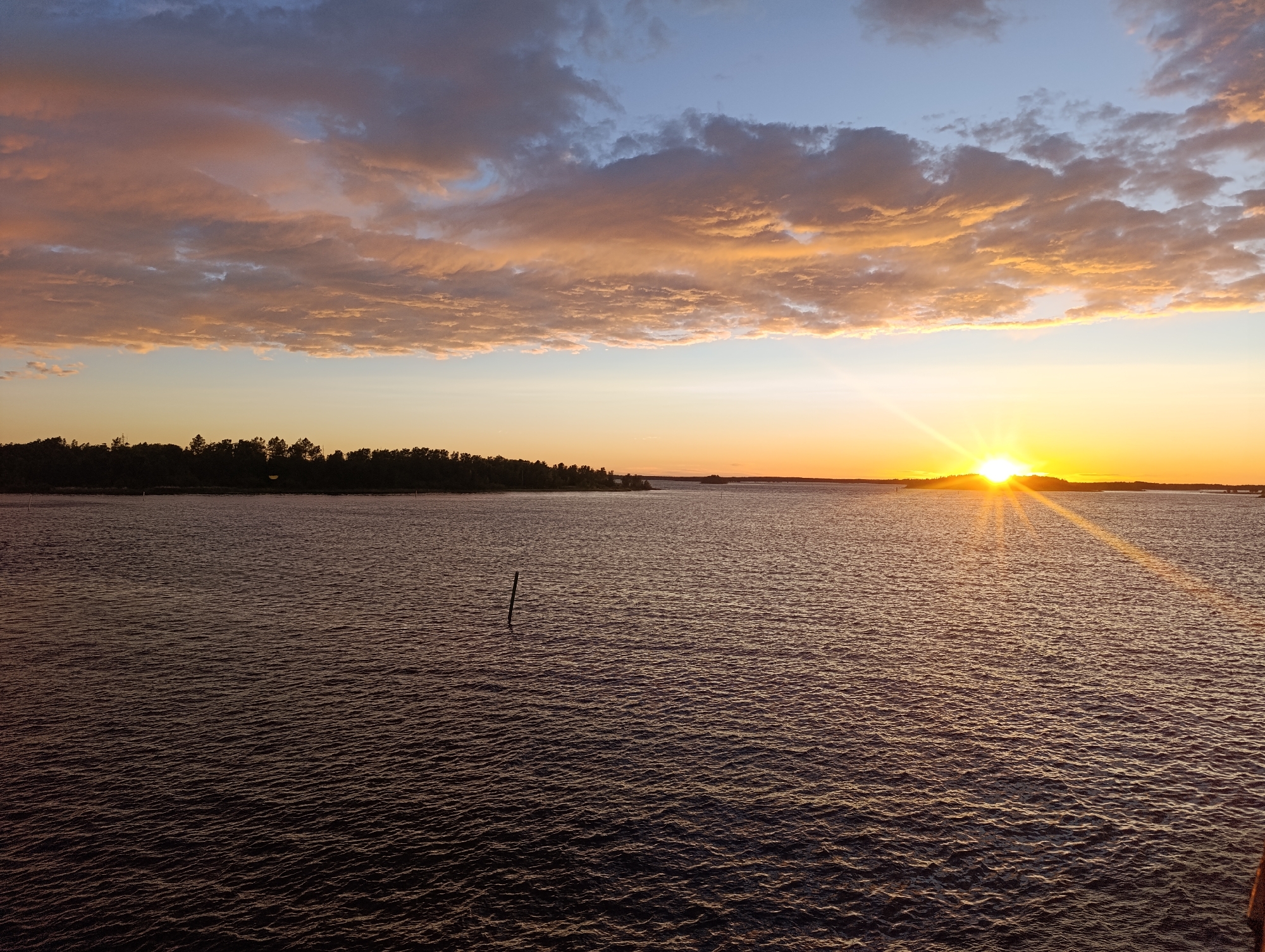 The image captures a sunset over the Baltic Sea, likely near Jakobstad, Finland. The sky is a gradient of colors, transitioning from a pale yellow and orange near the horizon to a muted blue and purple higher up. Wispy clouds are illuminated by the setting sun, displaying hues of orange and pink. The sun itself is a bright orb, casting rays of light across the water.
The sea occupies the majority of the foreground and middle ground, its surface rippled with small waves. The water appears dark, reflecting the darkening sky and the silhouette of the land. On the horizon, a dark, tree-lined landmass is visible, indicating a coastline. A single, dark pole or post juts out of the water in the mid-left of the image. The overall impression is one of tranquility and natural beauty at the end of the day.
