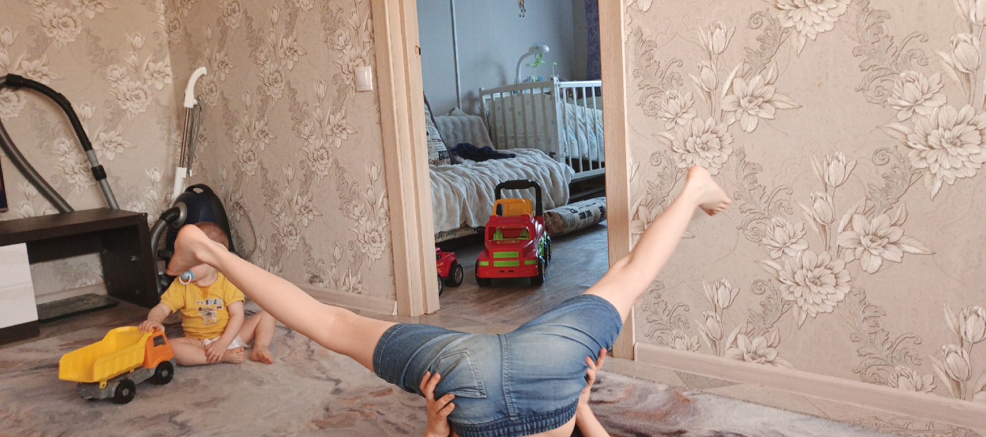 A child is seated on a carpeted floor in a room, next to a yellow toy dump truck, wearing a yellow t-shirt and diaper. A woman in denim shorts is practicing a yoga pose with her legs spread apart and raised, back arched, and torso on the floor. In the background, through an open doorway, a crib and a red toy truck are visible in another room. A dark brown shelf unit, a vacuum cleaner, and floral patterned wallpaper can be seen in the room where the child and woman are located. The location is Chaikovsky, Russia.