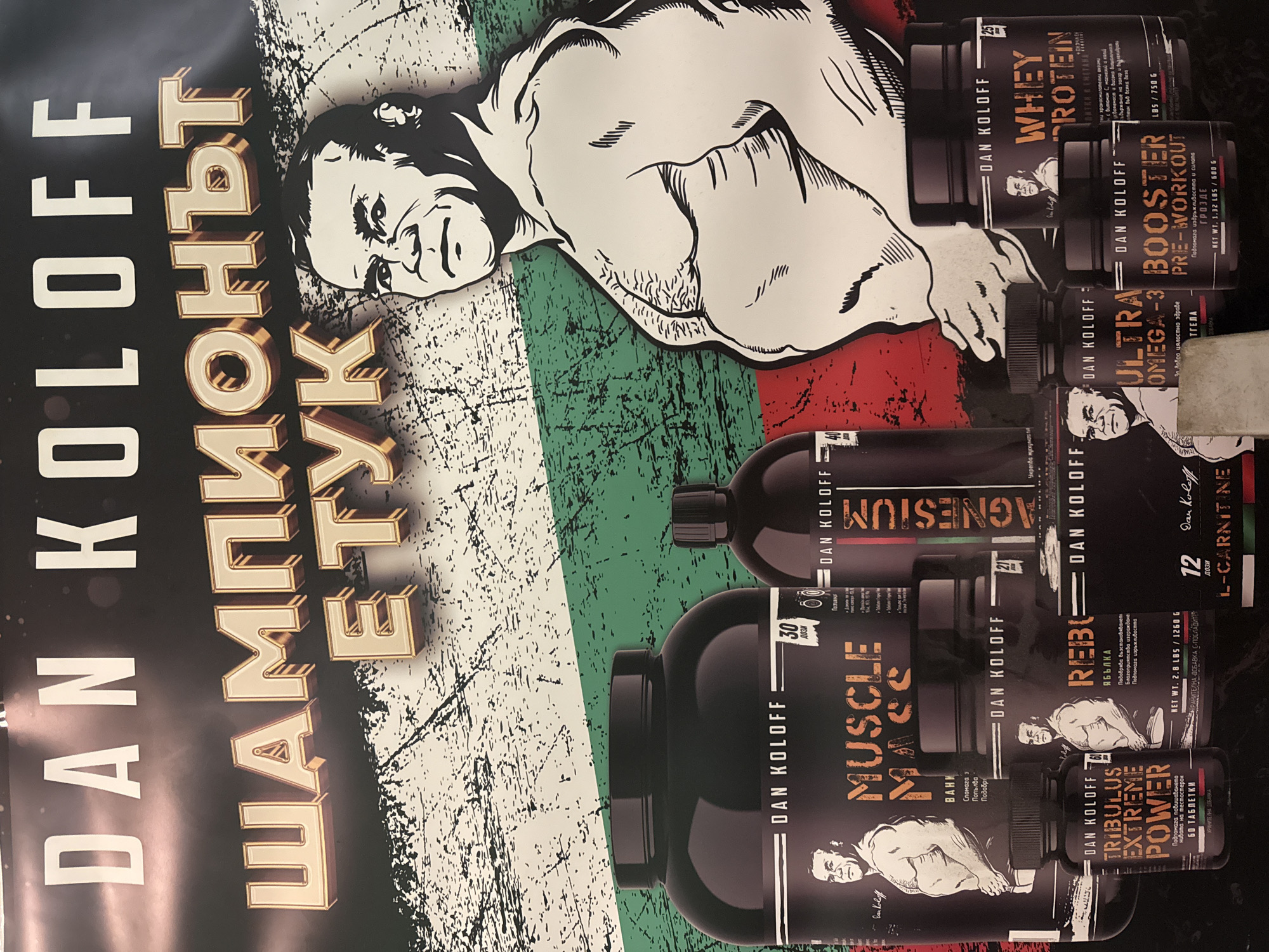 The image displays a promotional poster for Dan Koloff nutritional supplements, seemingly located in Varna, Bulgaria. The poster features a black and white cartoon-style drawing of Dan Koloff himself, overlaid on what appears to be the Bulgarian flag with a distressed or aged texture. A collection of black containers of the supplements are vertically arranged and oriented sideways next to the illustration of Dan Koloff. The supplements include various products such as whey protein, booster pre-workout, omega-3, magnesium, muscle mass, rebuila, tribulus extreme power, and l-carnitine. Text in Cyrillic alphabet is displayed on the left side of the image. The product labels and text on the poster are written in both English and Bulgarian, further suggesting the location of the poster in Bulgaria.