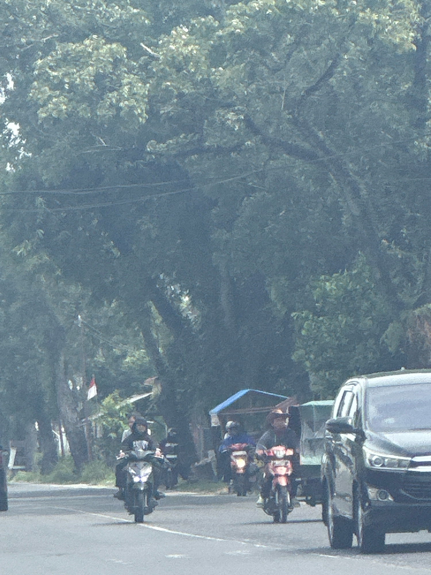 The image depicts a street scene in the City of Binjai, Indonesia, with a focus on vehicular traffic and the surrounding environment. In the foreground, a black SUV is on the right side of the frame. To the left of the SUV and slightly behind it, two motorcycles are visible. The motorcycle closest to the camera is being ridden by a person wearing a dark hooded sweatshirt. Further to the left and slightly behind this motorcycle, another motorcycle is moving, with its rider mostly obscured. In the background, several trees line the street, creating a canopy over the road. There are also some small structures and what appears to be a flag with red and white colors, possibly the Indonesian flag, visible in the middle ground to the left. The overall atmosphere suggests a typical day in a Southeast Asian city with moderate traffic.