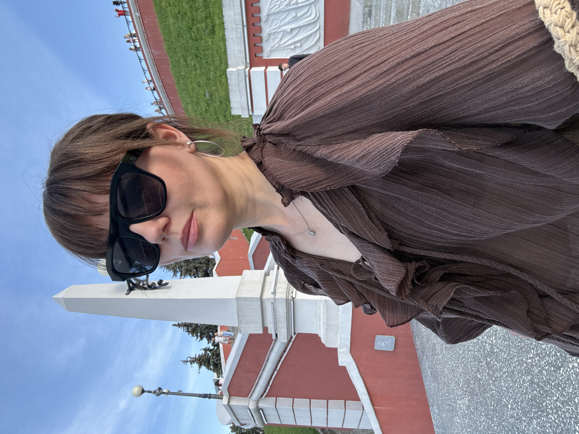 A woman, facing the camera and wearing large, dark sunglasses, is captured from the chest up in a portrait orientation. She has brown hair and is wearing a brown, crinkled blouse with ruffled sleeves and a delicate silver necklace. Her lips are slightly parted, and she appears to be looking at the camera with a neutral or slightly smiling expression.
In the background, a white obelisk stands prominently, with a dark, coiled metal sculpture near its top. Behind the obelisk, there's a large, red building with white architectural details and a row of green trees. The sky above is a clear, bright blue, suggesting a sunny day.
Further in the background, a grassy area slopes upwards, bordered by a railing. On this elevated area, several people are visible, some standing and some in a seated position, suggesting they are observing something. The overall scene appears to be outdoors in a public space, possibly a historical or memorial site. The lighting suggests it is daytime, likely late morning or afternoon. The image is a selfie, indicated by the perspective and framing.