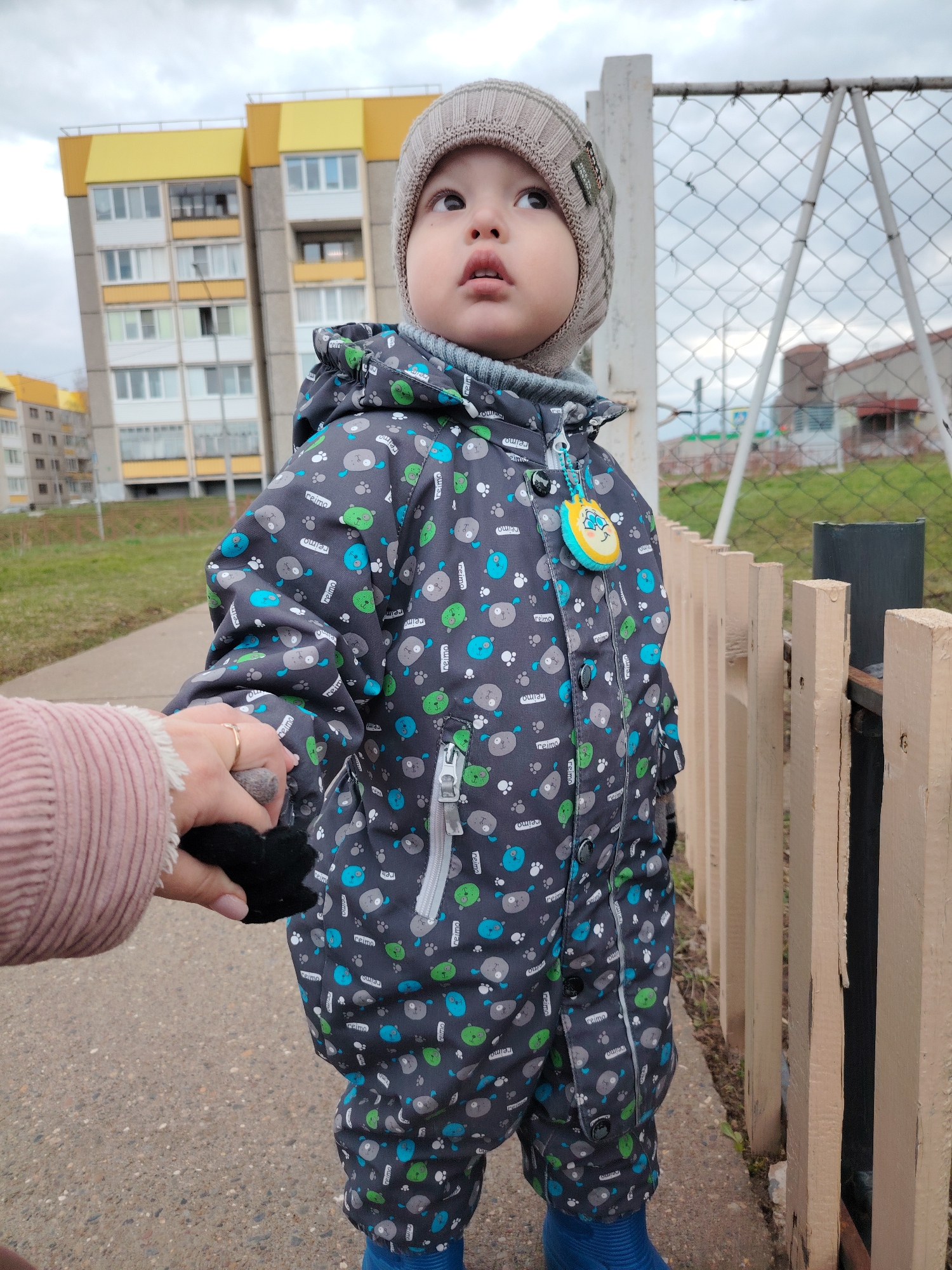 A young child, dressed in a patterned winter jacket and a knitted hat, stands on a paved pathway outdoors. The child is holding the hand of an adult, visible only as a pink-sleeved arm and hand clutching a small, black object. The background features a multi-story residential building with yellow and gray facades, a metal chain-link fence, and a light-colored wooden fence. The sky is overcast. The OCR detects the word "reimo" repeatedly on the child's jacket. The location is identified as the city of Markovskiy, Russia.