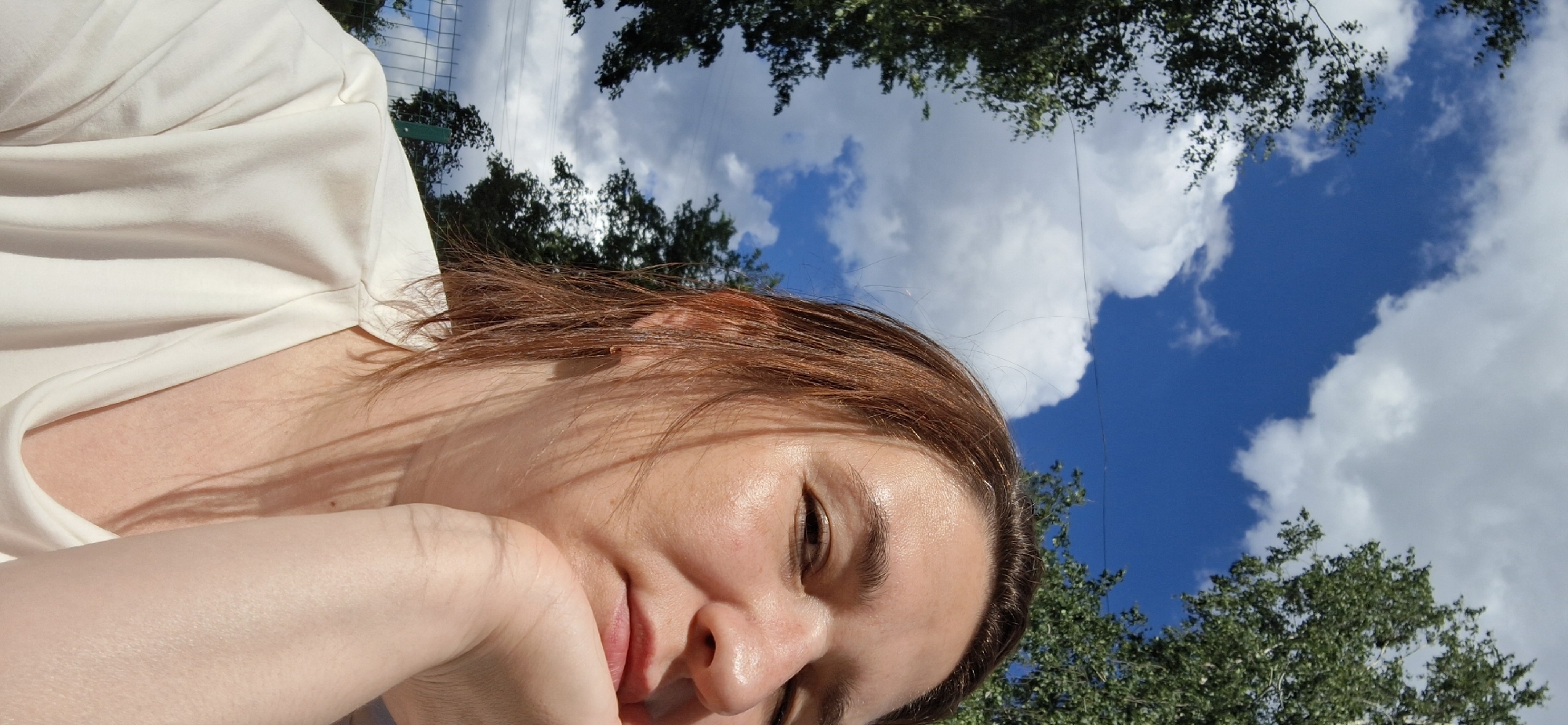 A close-up portrait shows the right side of a woman's face as she rests her chin on her hand. She has auburn hair, fair skin, and is wearing a white shirt. Behind her, a bright blue sky is filled with white clouds, and green trees frame the upper part of the image. A fence is visible in the lower left corner. The scene appears to be outdoors in the city of Chaikovsky, Russia.