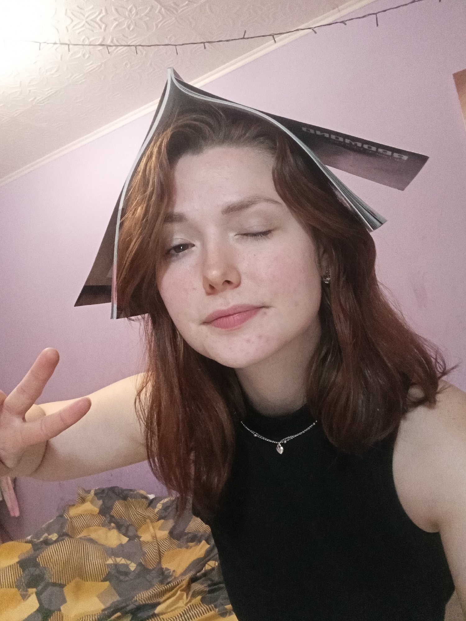 A person with auburn hair is indoors, wearing a black top and a silver necklace with a heart-shaped pendant. A magazine rests open on their head, forming a pointed hat shape. The person is winking, with their left eye closed and right eye open. They are holding up their left hand, forming a peace sign. The background consists of a light purple wall with a textured ceiling and a decorative string of lights. A patterned blanket or bedspread is visible in the lower-left corner. The location in the city is unknown.