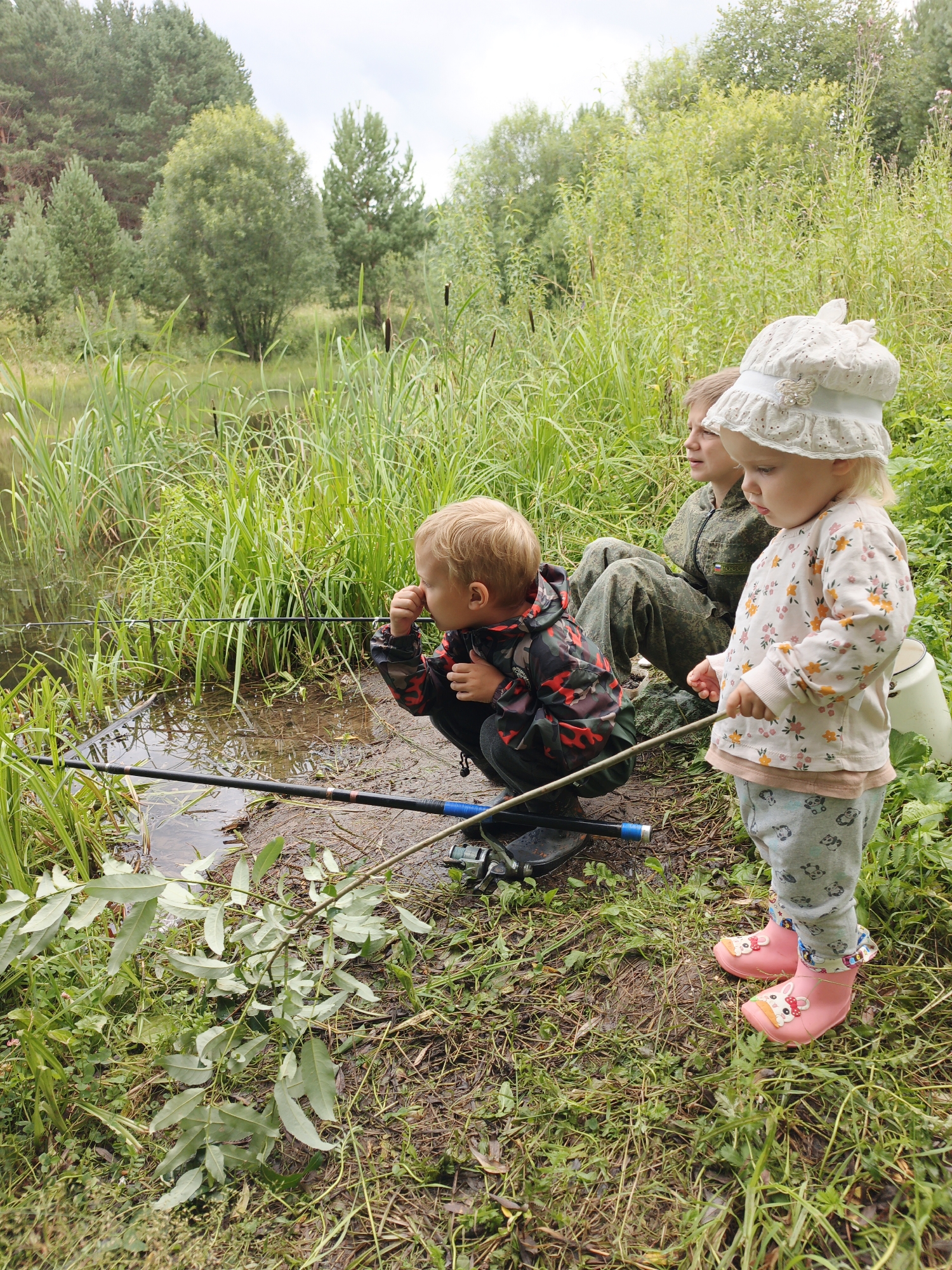 Three children are fishing by a body of water on a cloudy day. The location is the village of Medvedyovka, Russia. In the foreground, a young boy in a camouflage jacket and black boots is crouched by the water's edge, holding a fishing rod. To his right, a toddler girl in a floral shirt, patterned leggings, and pink rubber boots stands with a fishing rod. Behind them, a slightly older boy in camouflage clothing sits in the tall grass. The background is filled with lush green foliage, including trees and reeds, and the water reflects the overcast sky. The ground is muddy and covered in grass and fallen leaves. Two fishing rods are visible, one in the boy's hand and another lying on the ground.