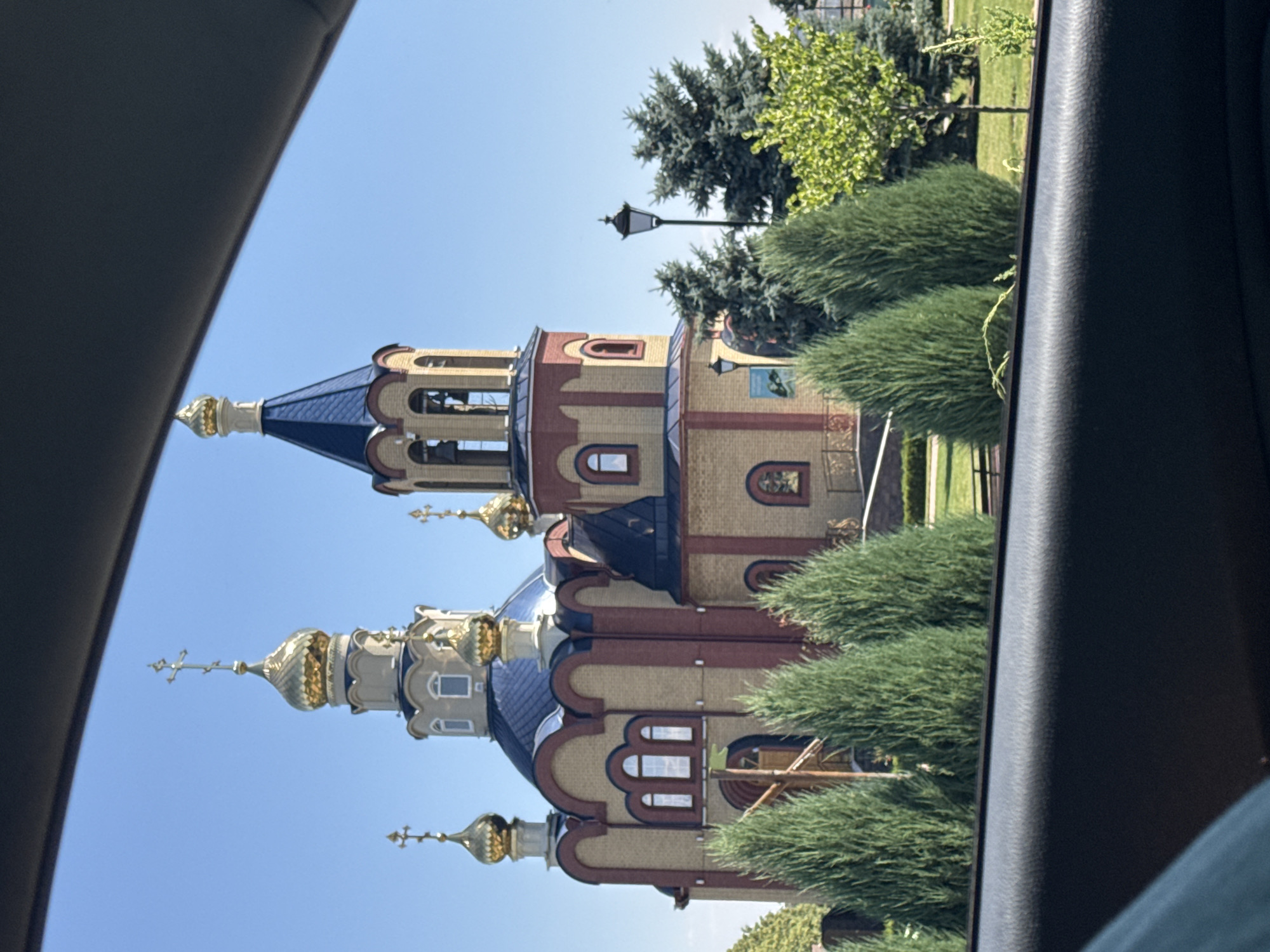 The image shows an Eastern Orthodox church with golden domes, a blue roof, and tan and red brick walls. The church is situated in a well-maintained outdoor setting with a clear blue sky, green grass, and several evergreen trees. A lamppost is visible to the right of the church. The foreground of the image is dominated by the interior of a vehicle, with a dark-colored dashboard or armrest in the lower portion. This suggests the photo was taken from inside a car looking out at the church. The church appears to be located in the village of Voronovo, Russia.