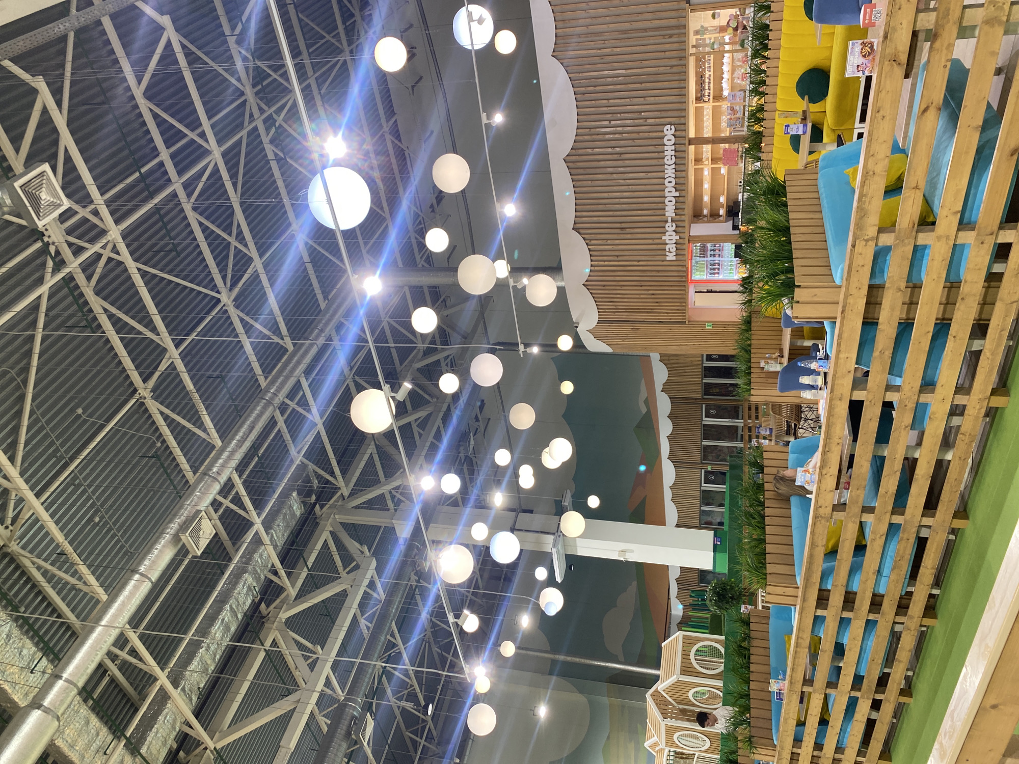 The image depicts an interior space, possibly a shopping mall or entertainment center in Perm, Russia, based on the Russian text visible ("кафе-мороженое" - cafe-ice cream). The space has a high, exposed ceiling with a network of metal beams and pipes. Multiple spherical lights hang from the ceiling, creating a bright ambiance.
In the foreground, there's a seating area with wooden fences or partitions. The seating features blue and yellow cushions. People are seated, engaging in activities such as reading or relaxing.
Beyond the seating area is a facade that includes a stylized landscape mural and a structure that resembles a playhouse. The space seems designed for families with children, given the presence of play areas.
The lighting is bright and somewhat harsh, creating a contrast between the ceiling structure and the more decorative elements of the interior design. The image provides a view of a relaxed environment within a larger, structured space.