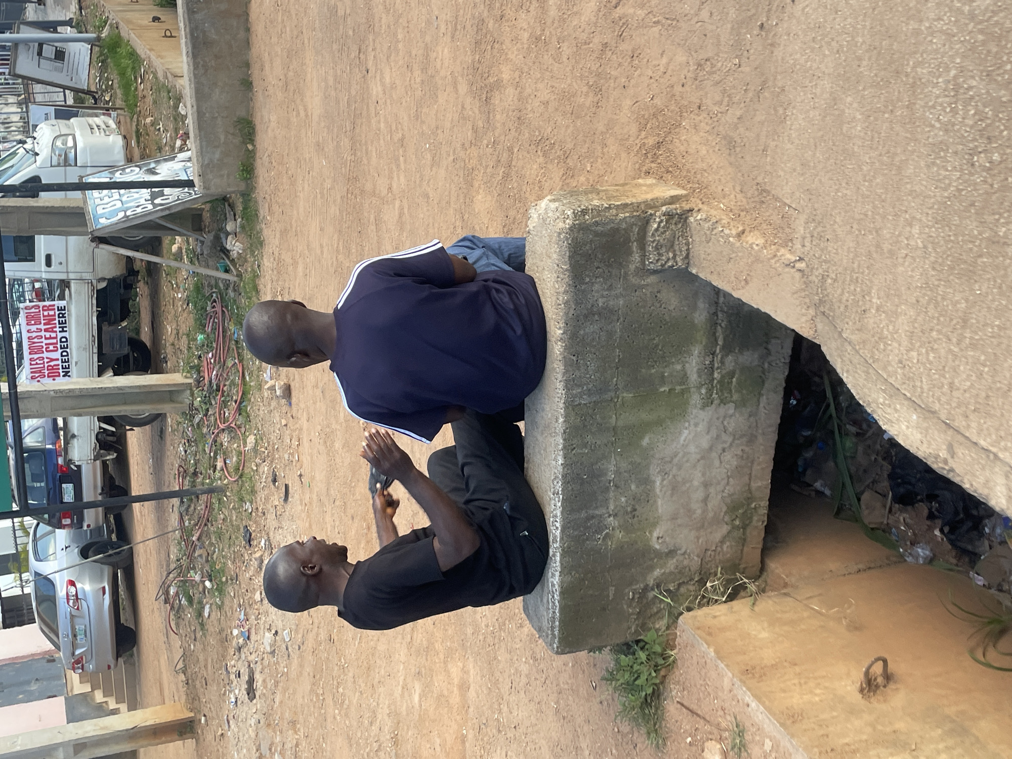 Two men are seated on a concrete structure with one man in a black t-shirt and black pants, and the other in a navy blue t-shirt and blue jeans. The man in black is holding a black object, possibly a phone, in his hands, and appears to be speaking. The man in navy blue is facing away from the camera. The concrete structure is positioned above a drainage ditch filled with refuse. In the background, a silver car and a white truck with signage are visible. The environment appears to be an urban setting in Jos, Nigeria, with unpaved ground and a building in the distance.