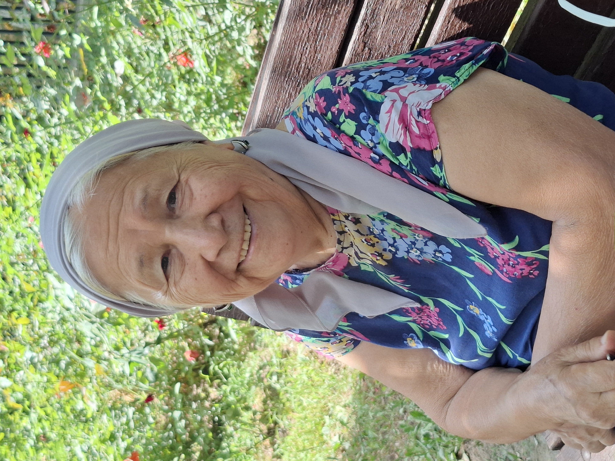 An elderly woman with a light grey headscarf and a blue floral dress with pink and white flowers is sitting on a wooden park bench outdoors. She is smiling and looking at the camera. The background is a blurred green foliage with some red flowers. The scene appears to be in a park or garden.