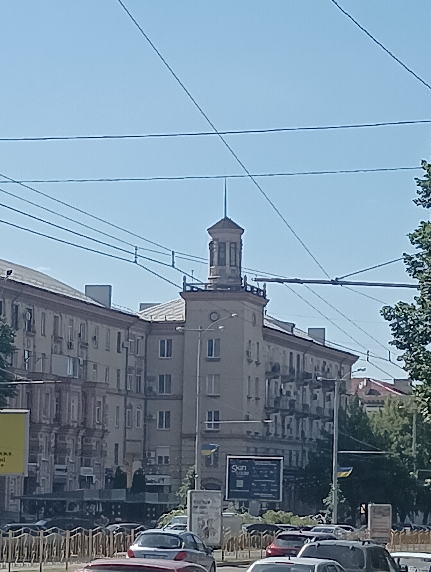The image shows a daytime exterior view of a city street with buildings and a clear blue sky. A prominent building with a tower is visible in the center, adorned with architectural details and topped with a spire. Power lines crisscross the sky above the street. To the left of the tower, a long apartment building with many windows and balconies stretches across the frame. On the right side, a tree with green foliage can be seen. The street level features a road with several cars, including a silver hatchback and a darker SUV, suggesting active traffic. A decorative metal fence runs along the sidewalk. Various advertisements and signs are posted on the buildings, one of which is a dark rectangular billboard with text and graphics. A Ukrainian flag is also visible, attached to a pole near one of the buildings. The overall scene depicts a typical urban environment on a sunny day.