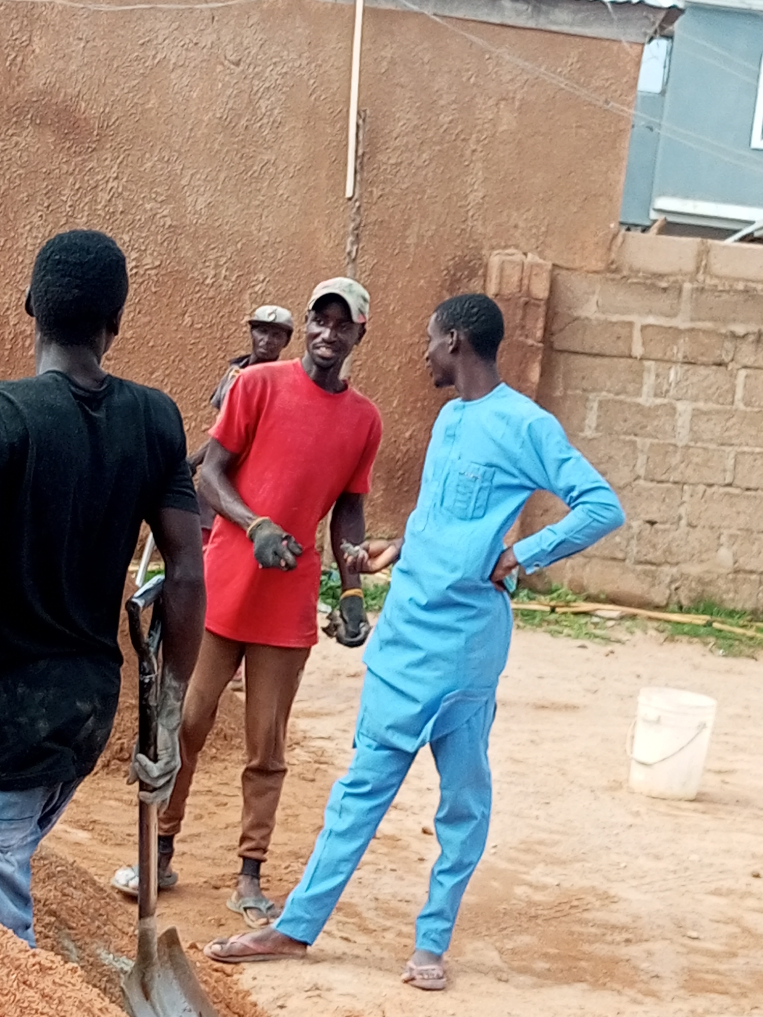 A group of men are working on a construction site in Tempure, Nigeria. The image shows three men in the foreground and one in the background. One man, wearing a black t-shirt, is facing away from the camera, holding a shovel. Another man, wearing a red t-shirt and a camouflage cap, is facing the camera and appears to be talking to the man in the blue attire. This third man, dressed in a blue kaftan, is also facing the camera and has his hand on his hip. A fourth man, wearing a cap and a red shirt, is visible in the background between the man in black and the man in red. The construction site is characterized by a brown textured wall, a brick wall, and a sandy ground. A white bucket is visible in the background.