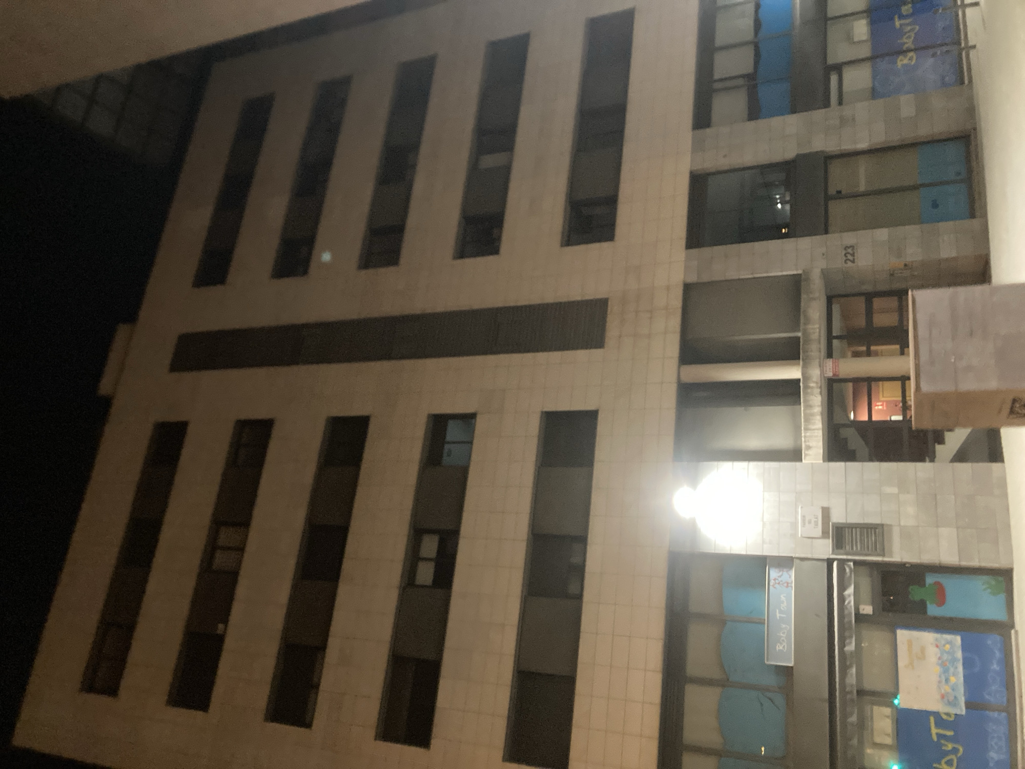 The image shows the exterior of a multi-story building at night. The building has a light-colored tiled facade with horizontal rows of dark windows. The ground floor appears to house commercial establishments, with signage visible including "Baby Town." The street in front of the building is paved, and there is a street light illuminating the scene. The overall environment suggests an urban setting, likely in Barcelona, Spain, given the style of architecture and the presence of Spanish text on some signs. There are no people clearly visible in the image.