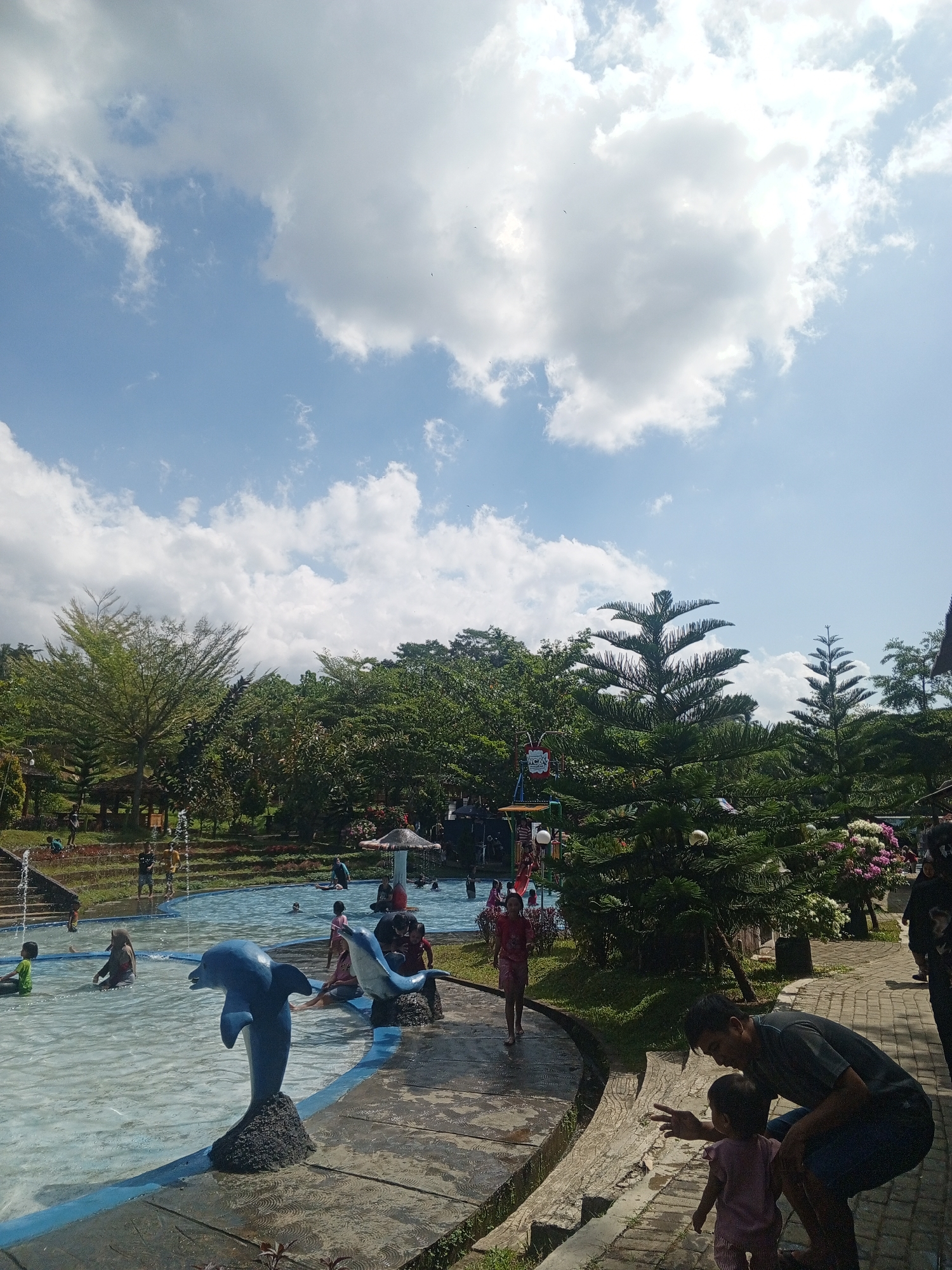 The image depicts a daytime scene at a public swimming pool with a water park area, likely located in Darsono, Indonesia, given the context. The sky is partly cloudy with bright sunlight. In the foreground, a blue dolphin statue stands on a dark rock base in the shallow end of the pool. To the right of the pool, a man is crouched down, interacting with a small child who is wearing a pink outfit and standing on a brick pathway. The pathway curves around the pool and is bordered by lush green grass and trees.
Further into the pool, several individuals, including children and adults, are seen swimming, wading, and playing. One child in a green shirt is visible on the left side of the pool. A larger blue dolphin statue is positioned in the water to the left of the man and child. In the background, more trees and vegetation surround the water park, with glimpses of structures and water features, suggesting a recreational park. The overall atmosphere appears to be one of leisure and family enjoyment on a sunny day.