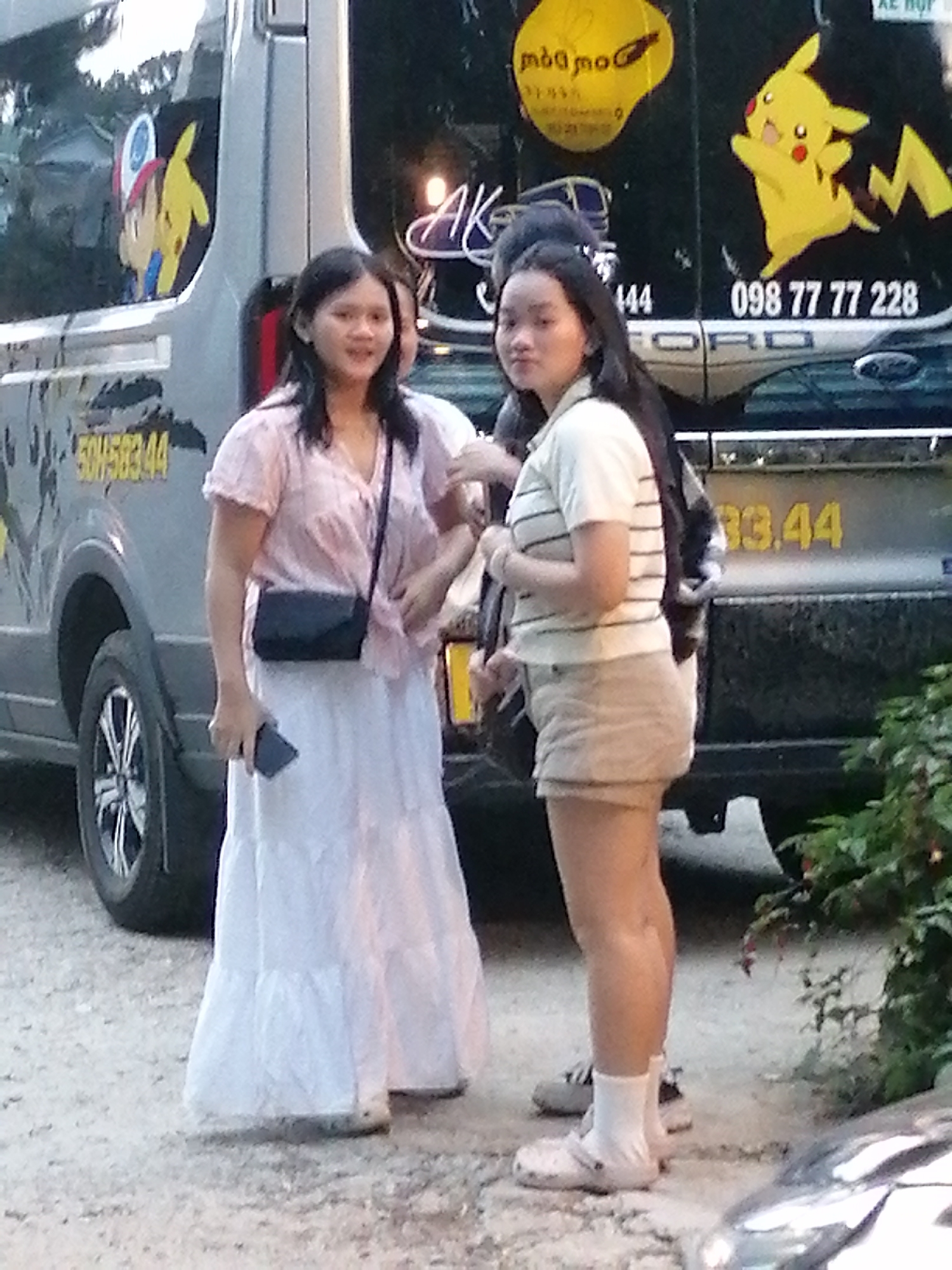 Two young women are standing near a silver van with cartoon decals on the back. The woman on the left wears a pink blouse and a long white skirt, carrying a black purse and a phone in her left hand. The woman on the right wears a striped t-shirt and light-colored shorts, with white socks and sandals. The van has text and images on its side, including "FORD" and a Pikachu sticker. The scene appears to be outdoors on a dirt or gravel surface, with some foliage visible on the right. This scene is located in Xuân Trường - Đà Lạt, Vietnam.