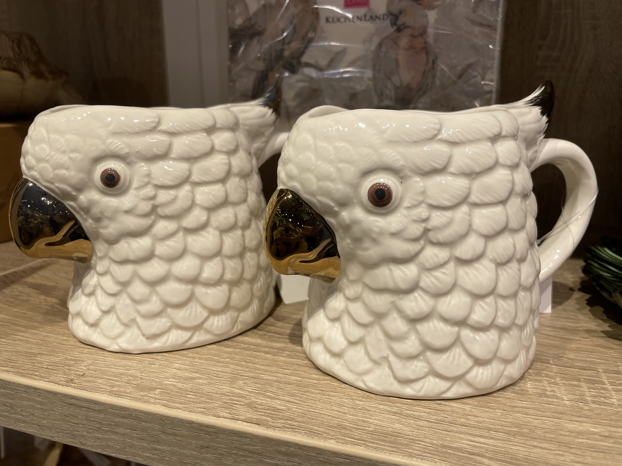 Two white ceramic mugs shaped like owl heads are displayed on a wooden shelf. The mugs have gold beaks and brown eyes. The owl's feathers are represented by raised textured scales. In the background, out of focus, is a plastic-wrapped item with text that appears to be in German, reading "KUCHENLAND". The scene is indoors, likely a retail environment, with the shelf appearing to be made of laminate. There is no indication of the city of Perm, Russia, in the image.