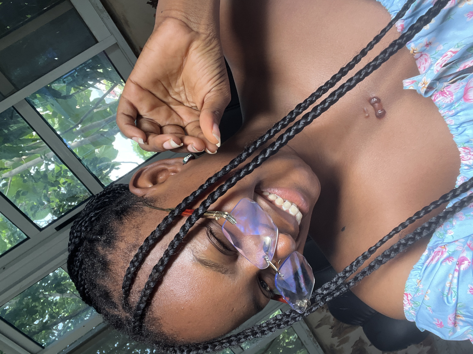 A young woman with dark skin is smiling in an indoor setting. She has braided hair, wears unique-shaped glasses with a purple tint, and has a light blue floral-patterned top. A navel piercing is visible on her stomach. Her hand is raised near her face. The background features a window with a view of greenery outside. There is no evidence suggesting a location in Adankolo, Nigeria.