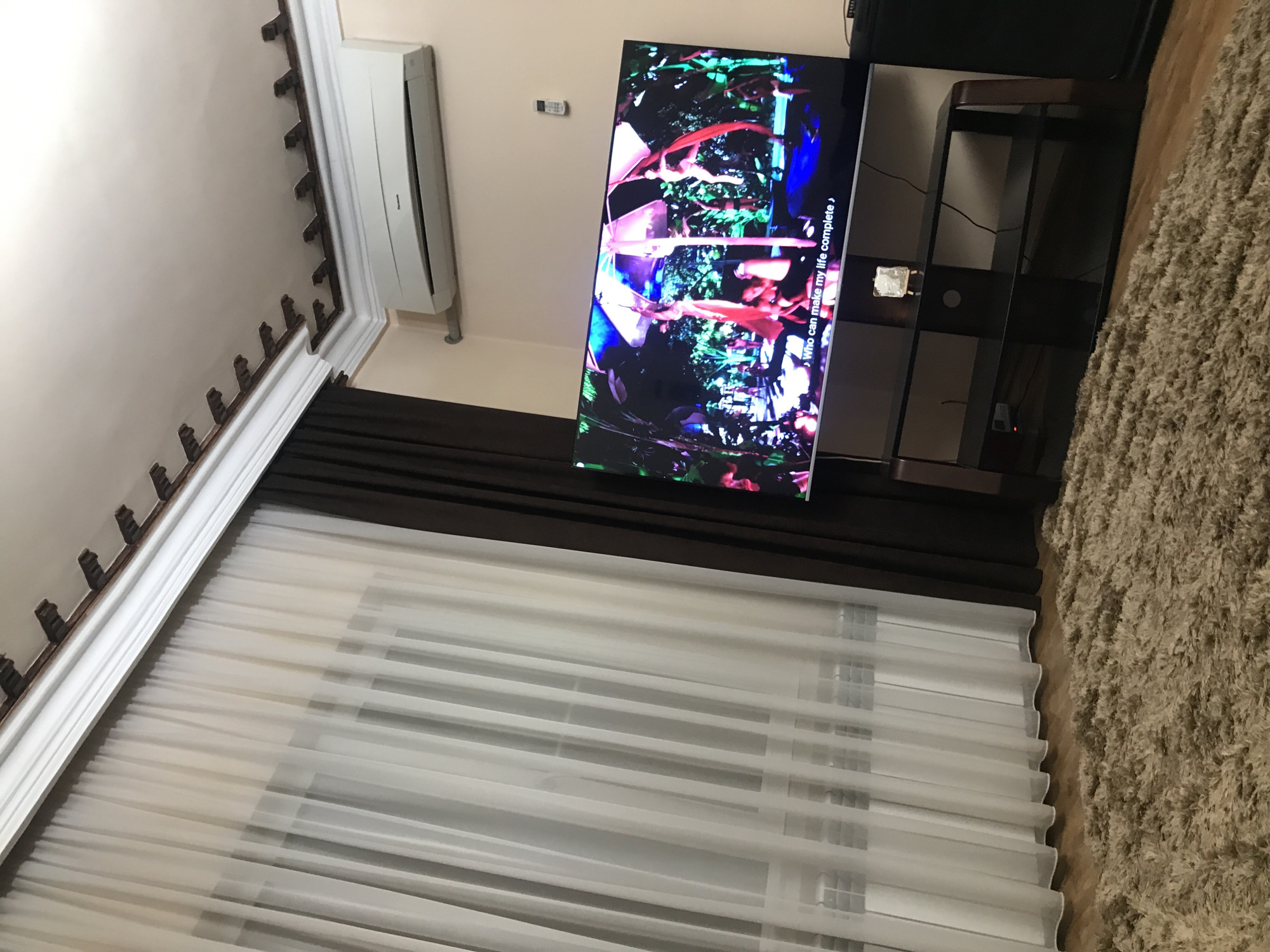 The image captures an interior setting. The focal point is a large television displaying an abstract, colorful scene. Adjacent to the TV is a black shelf unit, and a plush, beige carpet occupies the floor space.
The wall features an air conditioning unit and dark brown drapes. A window covered with sheer white curtains is visible below the drapes. A decorative ceiling trim is present along the top edge of the wall.
The photograph provides no information to determine if the location is in Yerevan, Armenia. There are no people visible in the image.