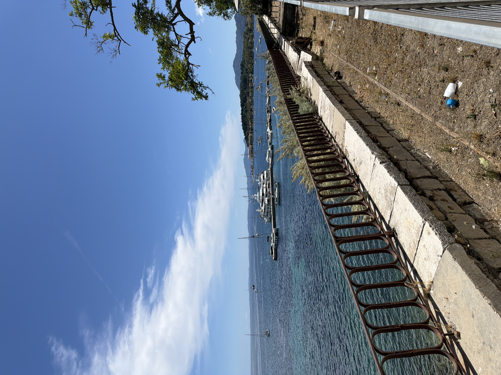 A vibrant, sun-drenched image presents a panoramic view of a tranquil coastal scene in Corfu, Greece. The perspective is elevated, looking down over a serene bay teeming with boats under a bright blue sky.
The left side of the frame features the expansive, clear blue water of the bay, displaying beautiful variations from light turquoise near the shore to deeper blues further out. Numerous boats, including several sailing yachts and motorboats, are visible throughout the bay, some moored near a pier or floating dock, others anchored in the open water. In the distance, a range of verdant mountains rises from the far coastline, completing the picturesque backdrop.
The upper portion of the image is dominated by a brilliant blue sky, adorned with scattered, wispy white clouds. Green leafy branches of trees extend into the frame from the top-left corner, providing a natural frame.
Along the right side of the image, a robust concrete embankment or retaining wall runs parallel to the water's edge. An old, rusty metal railing with an intricate, repeating oval design is affixed to this wall, providing a boundary between the land and the sea. Above the concrete wall, the ground appears dry and uncultivated, covered with sparse dry grass and small stones. Scattered debris, including a crumpled white item and a small blue object with a white top (possibly a buoy or bottle), are visible on the ground. Further back, a modern metal fence runs along the top of the embankment.
The scene captures a peaceful daytime atmosphere, characteristic of a clear day in a Mediterranean coastal area. No people or visible text are present in the image.