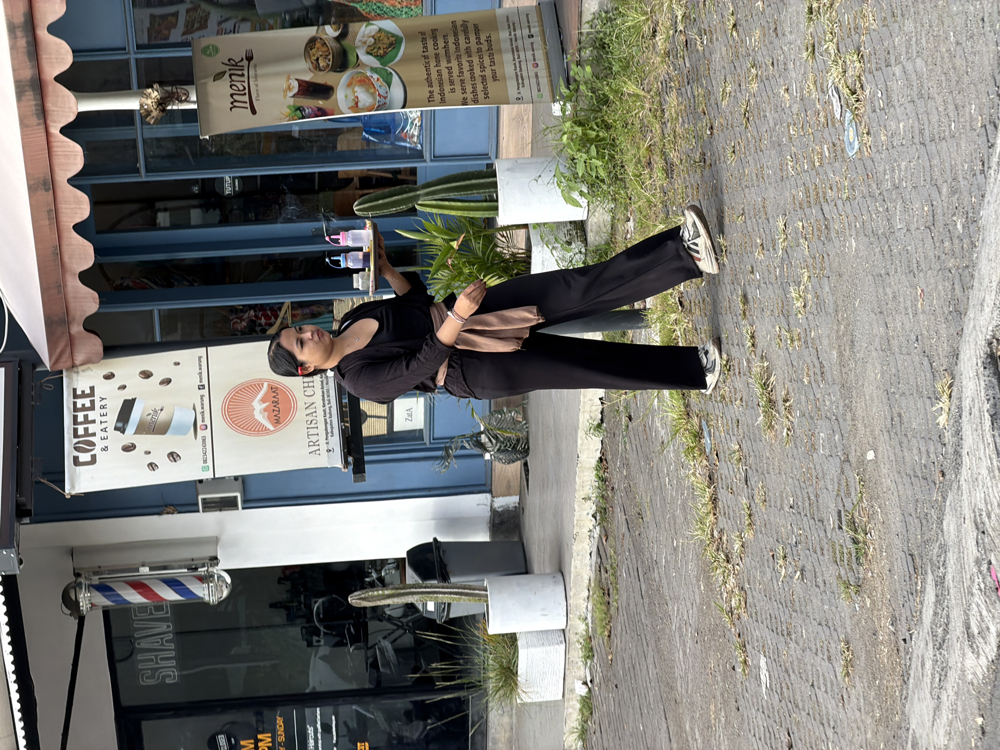 A woman wearing a black top, black wide-leg pants, and sneakers is walking on a paved walkway in front of shops. She is carrying a tray with two bottles and a small dish. A barber pole is visible on the left side of the frame, and banners advertising "Coffee & Eatery" and "Menik Flavor of Indonesia" are displayed. There are potted plants, including a tall cactus, near the entrance to the shops. The ground is paved with textured stones, and sparse grass grows between the stones. The scene appears to be in a commercial area in North Kuta, Indonesia.