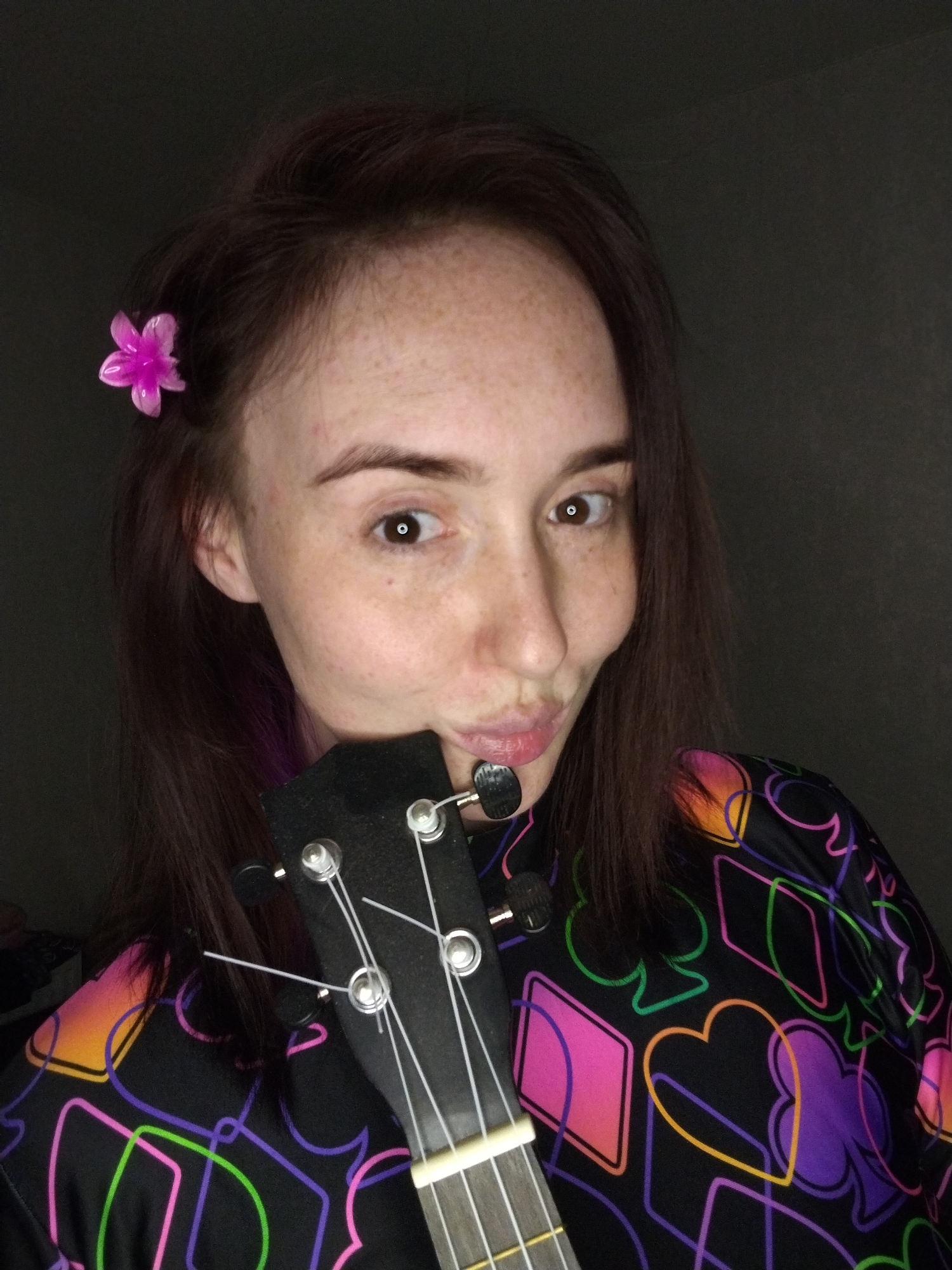 A woman with auburn hair and freckles poses close to the camera holding a ukulele. A pink flower is clipped in her hair. She is wearing a black top with neon-colored card suit symbols. The background is a dark gray wall. The location is not discernible and the city of Izhevsk, Russia cannot be determined from the provided image.