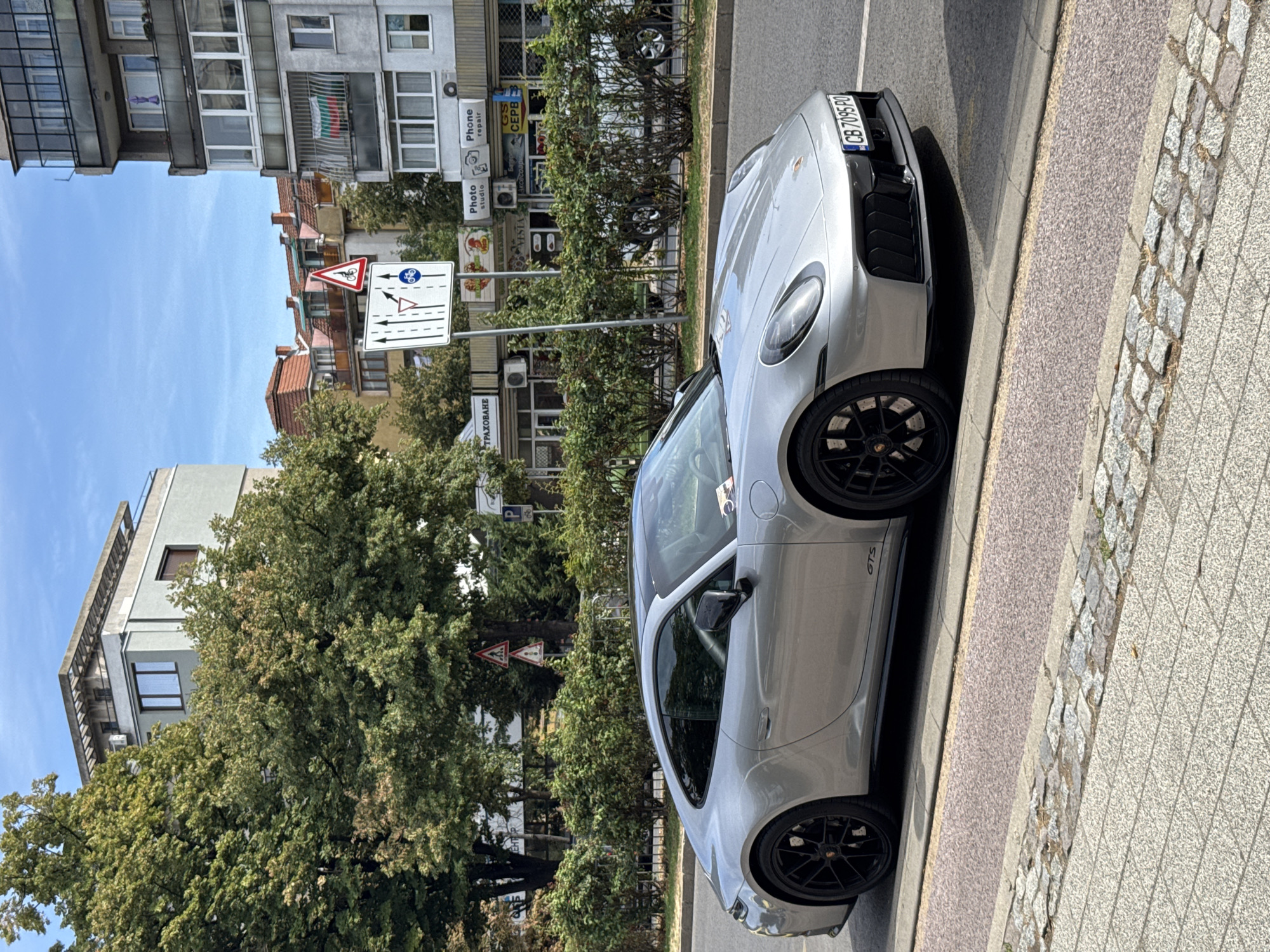 A silver Porsche 911 Carrera GTS is parked on a city street in Plovdiv, Bulgaria. The car is facing right, with its front passenger side towards the viewer. It is a sunny day, and the car is parked on the right side of the road, next to a sidewalk. The street is lined with trees and buildings. In the background, there are several buildings, including a multi-story residential building with balconies and a different building with a flat roof. Traffic signs and shop signs are visible on the buildings. The car's license plate is visible and reads "CB 7095 PO". The car has black wheels and a black roof. The overall scene depicts a typical urban street in Plovdiv on a clear day.