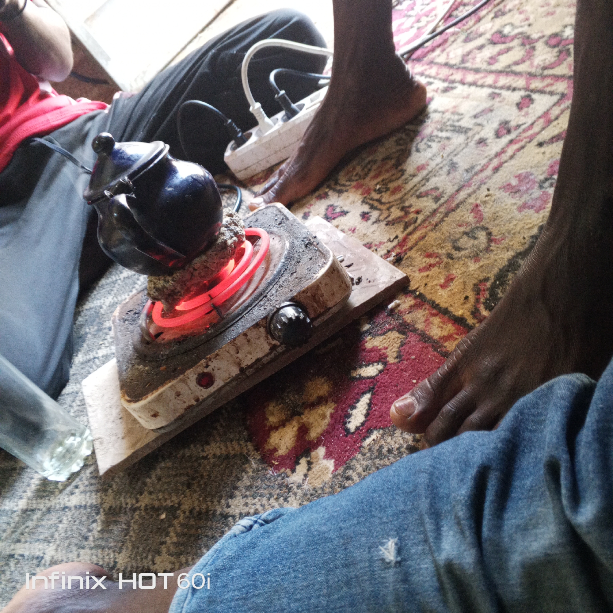 The image shows a dangerous setup with a blue teapot sitting on a hot electric stovetop burner. The burner is plugged into a power strip with other electrical cords. The scene is indoors, on a patterned rug. A person's bare foot is near the power strip and another person in blue jeans is sitting nearby. The picture was taken with an Infinix HOT 60i phone. Location is in the city of Unknown, Unknown.