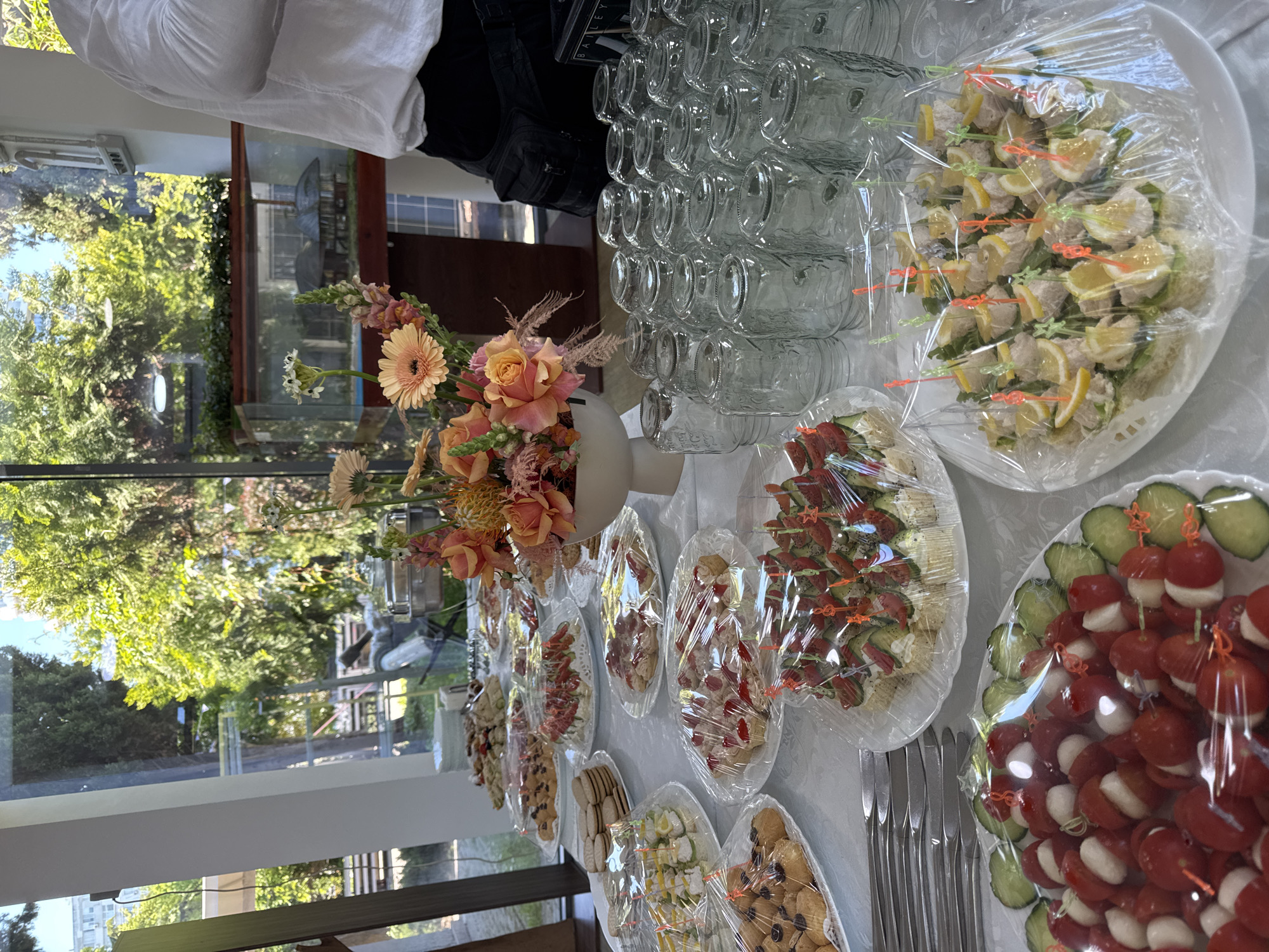 Here's a detailed description of the image:
**Overall Scene:**
The image shows a buffet table laden with food and refreshments, likely prepared for an event or gathering. The scene is indoors, close to a window with a view of trees and possibly a cityscape.
**People:**
A person wearing a white shirt and dark trousers is partially visible at the top of the frame, possibly a caterer or server.
**Subjects & Objects:**
* **Food & Drink:** The table is covered with multiple plates of hors d'oeuvres or canapés. The food is covered with plastic wrap. Items include:
* Stacks of clear glass cups.
* Sandwich skewers with lemon wedges.
* Skewers with tomato and cheese with cucumber.
* Other skewers featuring variations of meat, cheese, and vegetables.
* Cookies or small pastries.
* **Decor:** A white vase holds a floral arrangement featuring orange roses, gerbera daisies, and other foliage.
* **Utensils:** A stack of knives is present near the plates of food.
* **Window View:** The window reveals greenery, trees, and possibly a cityscape beyond.
**Actions & Interactions:**
The scene is static, suggesting preparation for an event. There is no visible interaction occurring at the moment the photo was taken.
**Environment:**
The location appears to be indoors, close to a window. Light is bright. The presence of prepared food indicates a catered event.
**Location:**
It is difficult to confirm the location in Burgas, Bulgaria without additional information.