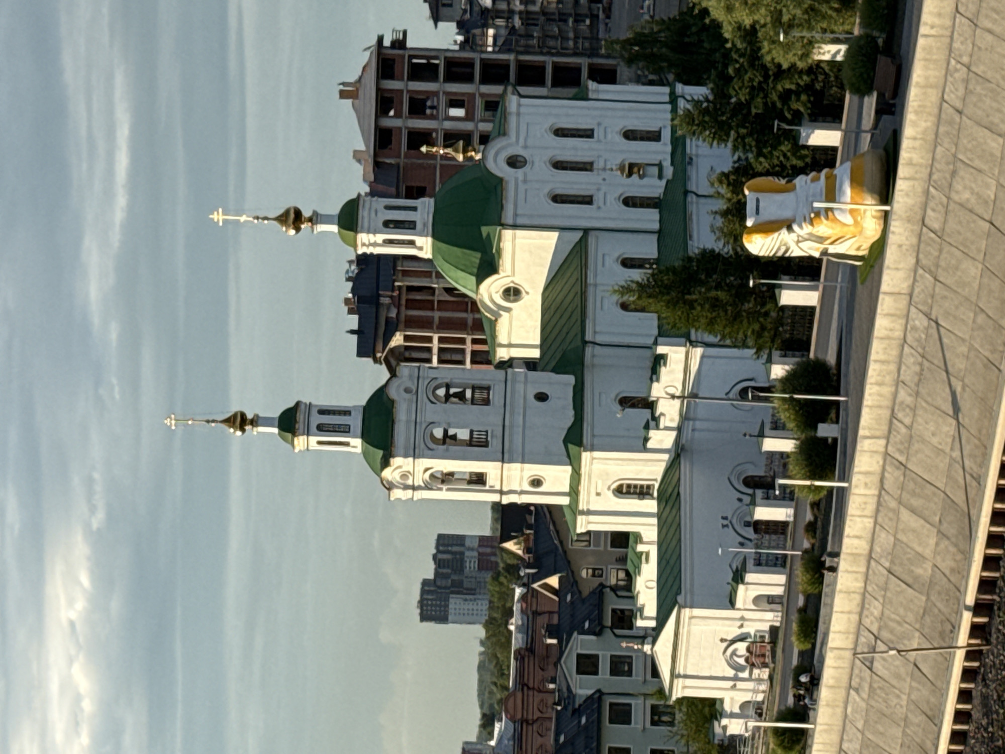 This is an aerial, medium shot of the Alexander Nevsky Cathedral in Tyumen, Russia, with the Tura River embankment in the foreground. The cathedral, a prominent white building with green domes and gold crosses, is the central subject. To the right of the cathedral, a large, yellow and white sculpture resembling a sneaker is positioned on the embankment. The embankment itself is made of concrete slabs and is bordered by a low wall. Several people are visible walking and sitting on the embankment. Behind the cathedral, residential buildings and construction sites are visible, suggesting an urban environment. The sky is partly cloudy. The lighting suggests it is either early morning or late afternoon.