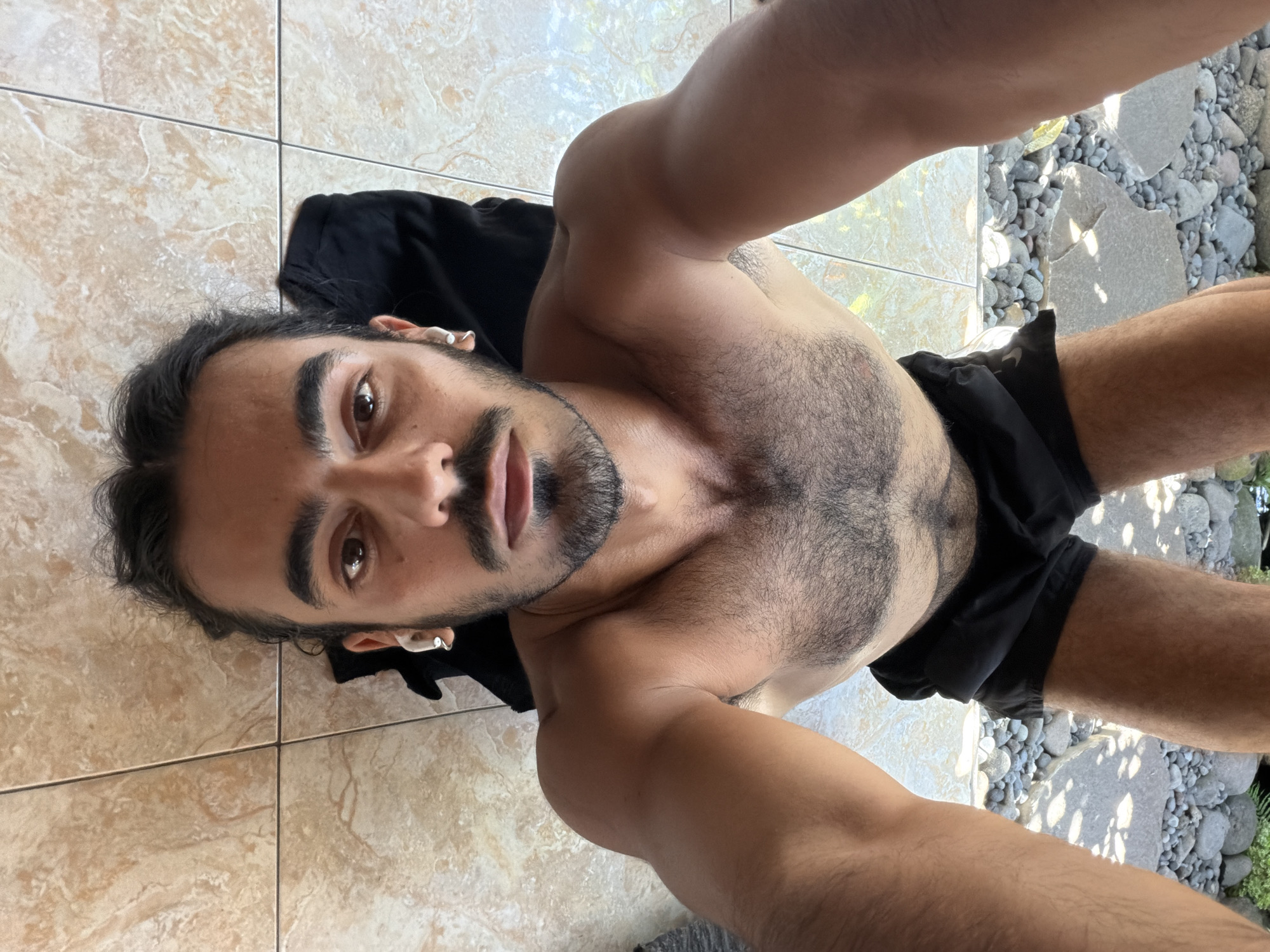 A shirtless man with dark hair and a mustache is taking a selfie. He is wearing black shorts and standing on a tiled floor that transitions to an outdoor area with small, smooth rocks. The photo was taken in North Kuta, Indonesia.