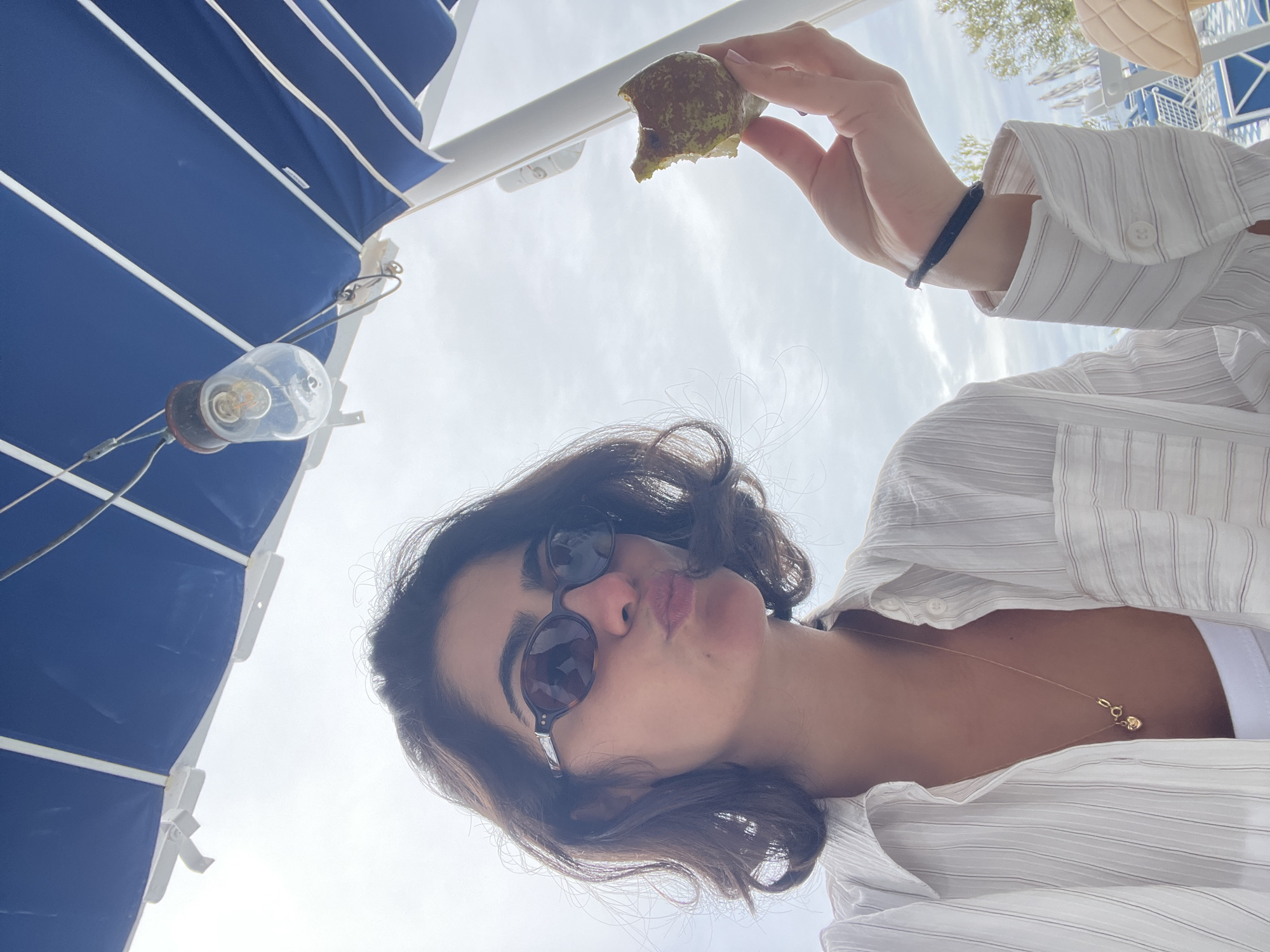 The image shows a woman holding a partially eaten piece of fruit, possibly a fig, above her. She is wearing sunglasses and a white, striped shirt. In the background, there is a blue and white striped awning and a lightbulb fixture. Given that the image is in Santa Eulària des Riu, Spain, the setting is likely an outdoor area, such as a terrace or patio of a restaurant or cafe.