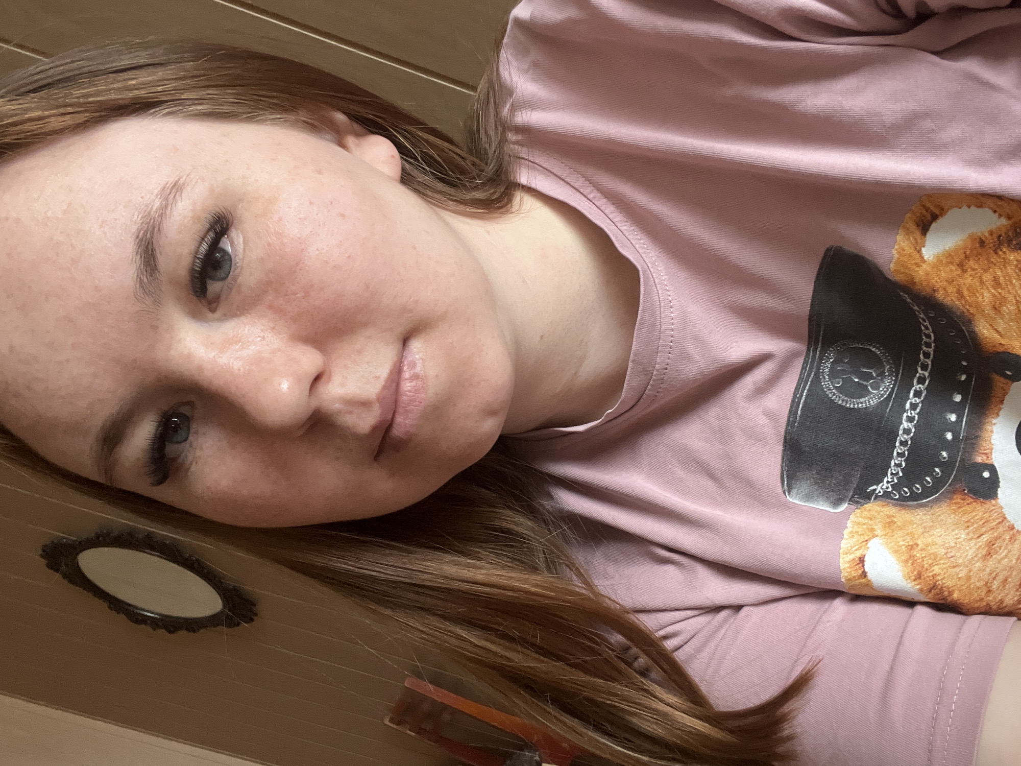 A young fair-skinned woman is lying down, facing upwards and slightly towards the left of the frame. She has red-brown hair, visible freckles on her face, and has long eyelashes. She is wearing a light pink long-sleeved shirt featuring a graphic of a teddy bear wearing a black hat with chains.
A small, decorative oval mirror is partially visible at the left edge of the image. There is wood paneling or a wooden surface visible in the background. A reddish-brown object is also visible near the bottom of the frame, partially obscured by her hair.
There is no information available in the image that would indicate that the location is the city of Chaikovsky, Russia.