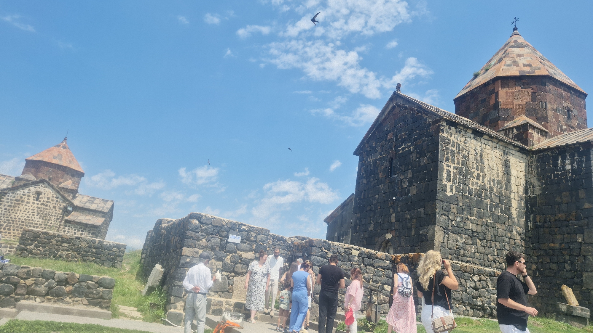 The image depicts the Sevanavank monastery complex, a historic religious site located on a peninsula overlooking the city of Sevan, Armenia. The architecture features dark, rough-hewn stone buildings with conical, reddish-brown tiled roofs, topped with crosses. Several people are visible, appearing to be tourists or visitors, some standing in small groups, others walking or observing the surroundings. The sky is a clear blue with scattered white clouds, and at least three birds are in flight. The ground is covered with grass and stone pathways and walls.