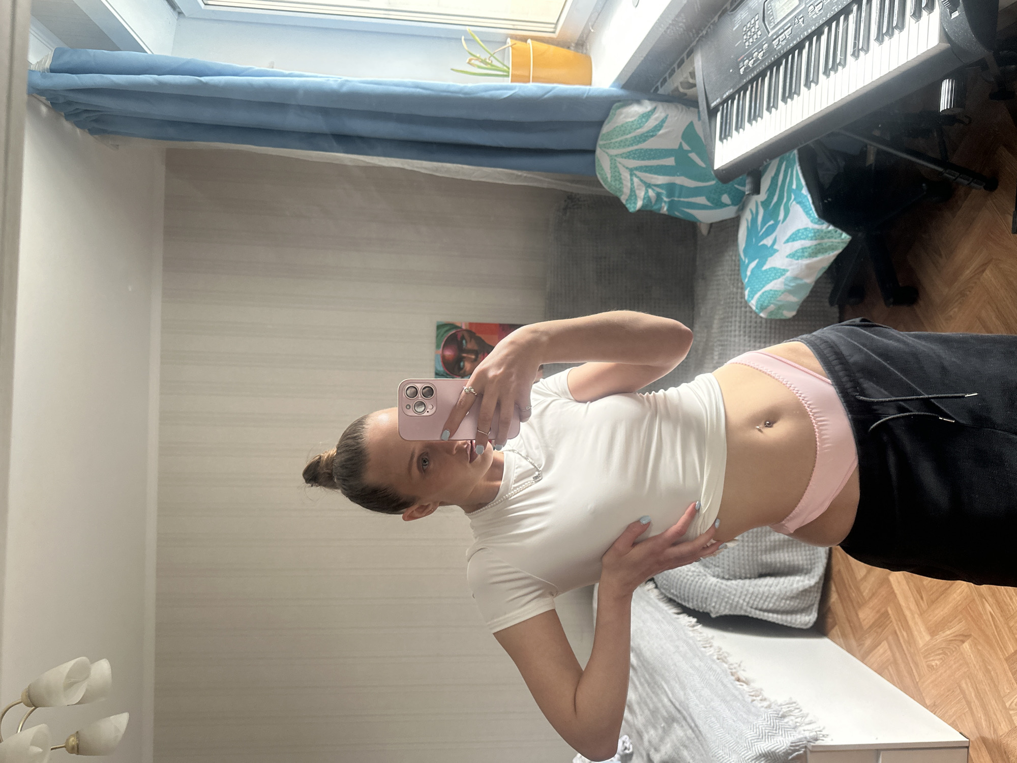 The media file shows a young woman taking a mirror selfie in what appears to be a bedroom. The woman is wearing a white t-shirt and pink underwear, with her belly button pierced. She has her hair pulled back in a bun and is holding a pink smartphone. In the background, a keyboard is visible, along with several decorative pillows with a tropical leaf pattern. The room appears to be lit by natural light coming from a window. The overall setting suggests a casual, indoor environment. The time of day and weather are not discernible from the image. No text is visible in the image.