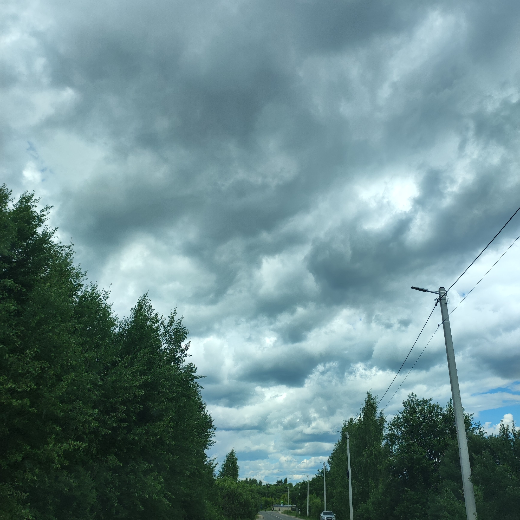 The media file captures a view of a rural road lined with dense green trees on either side, under a dramatic, overcast sky filled with heavy, gray clouds. In the foreground, to the left, is a thick canopy of lush green foliage from the trees. To the right, a concrete utility pole with a streetlamp and wires stands prominently, stretching upwards towards the sky. Further down the road, more trees and utility poles can be seen, with a faint hint of blue sky peeking through the clouds in the distance. A silver car is parked on the right side of the road near the utility pole. A small green road sign with a white symbol is visible in the distance on the left side of the road. The overall mood is somber, suggesting the imminence of rain or a storm. The scene appears to be a landscape shot of a road in Kineshma, Russia, likely during the daytime, given the bright clouds, though the heavy cloud cover creates a dusky atmosphere.