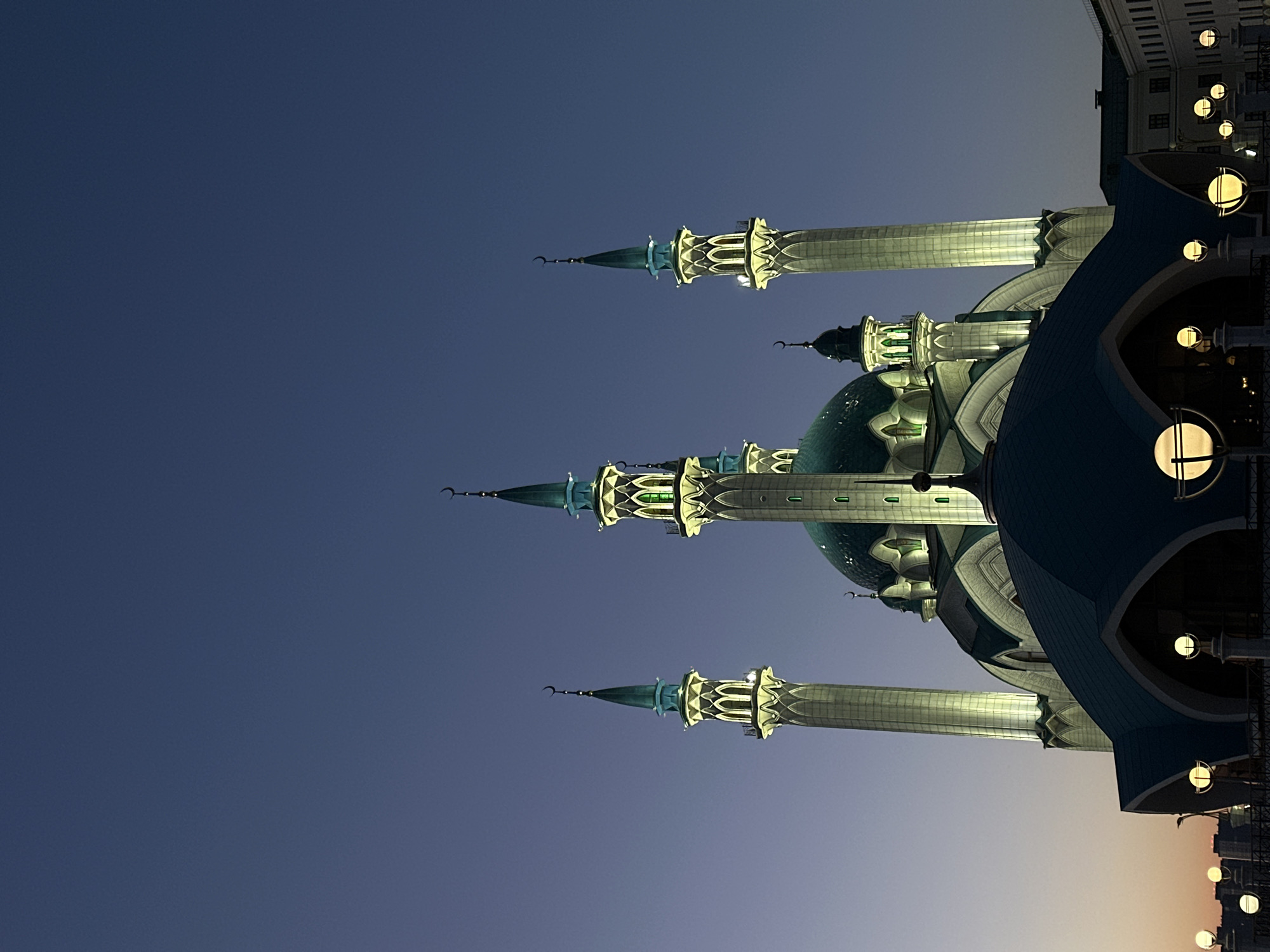 The image shows the Kul Sharif Mosque in Kazan, Russia at night. The mosque features multiple minarets and a blue dome, all illuminated. Street lights can be seen on the right side of the image, while the rest of the sky is a gradient of blue.