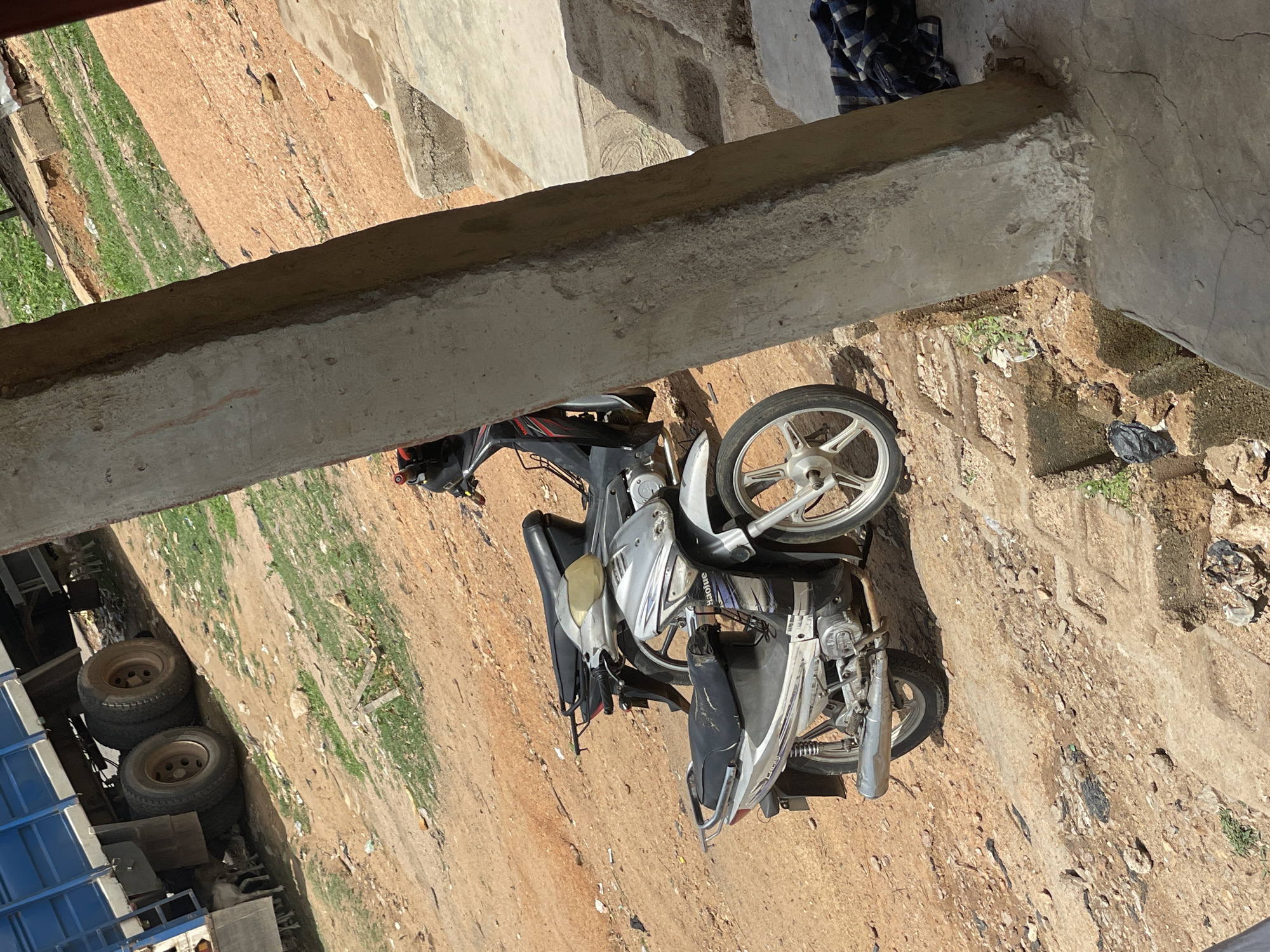 The image shows a parked motorcycle on a dirt ground in Mararaba, Nigeria. The motorcycle is silver and black, with a front wheel that has a distinct spoke pattern. Above the motorcycle, there is a concrete structure. A dirt path or area with some scattered vegetation runs adjacent to the ground where the motorcycle is parked. In the background, parts of a blue truck with visible wheels are seen. The setting appears to be an outdoor area.