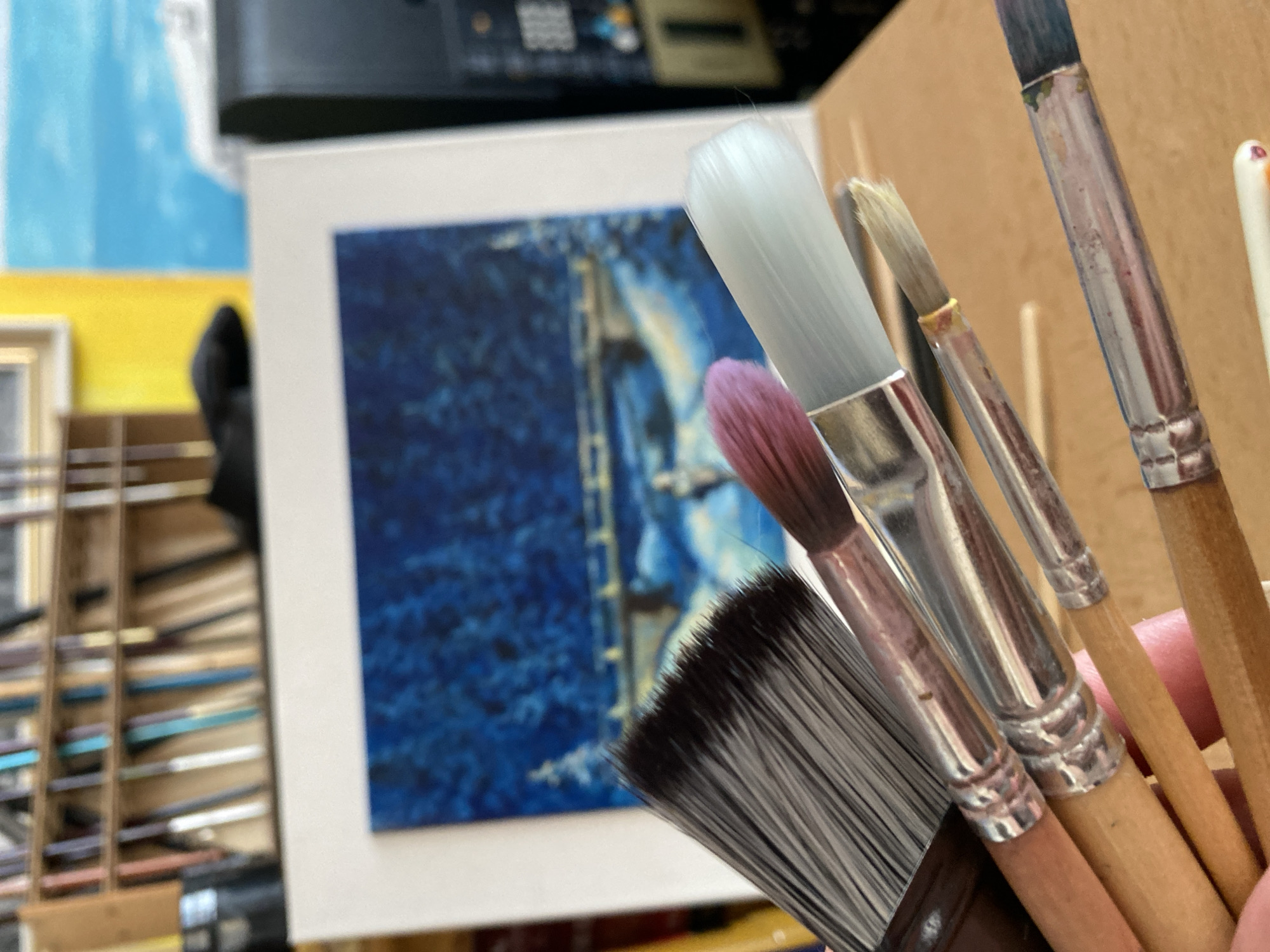 This is a close-up, eye-level, medium shot of several artist's paintbrushes held in a person's hand, foregrounded against an out-of-focus background that suggests an art studio. The brushes are the primary subject, with their wooden handles and metal ferrules clearly visible. The bristles of the brushes vary in size, color, and texture, with one large, dark grey brush dominating the left side of the frame. To the right of this, a smaller brush with pinkish-purple bristles is visible, followed by brushes with white, light brown, and dark blue bristles. Some of the brushes show signs of use, with paint residue visible on the bristles and ferrules.
In the background, a framed painting depicting a snowy landscape with a bridge and trees is partially visible. Above this, shelves filled with art supplies, including tubes of paint and more brushes, are arranged. The overall impression is one of a creative space, likely a personal art studio, where an artist is preparing to work or has just finished a painting session. The image was captured in the city of Bílina, Czechia, but the specific location within the city is not identifiable from the visual cues present.