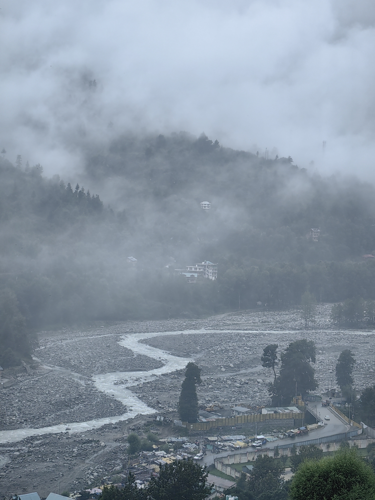The image shows a panoramic view of a mountainous landscape in Vashisht, India, shrouded in fog. A winding river, appearing milky white, flows through a valley filled with grey stones and sparse vegetation. On the slopes of the mountains, evergreen trees are visible, partially obscured by the mist. Several white, multi-story buildings are scattered across the hillsides, suggesting a settlement. In the foreground, a road curves through a built-up area with more buildings, some appearing to be commercial establishments or small residences. Vehicles, including cars and what appear to be construction vehicles, are present on and around the road. The overall atmosphere is somber and diffused due to the heavy fog and overcast sky.
