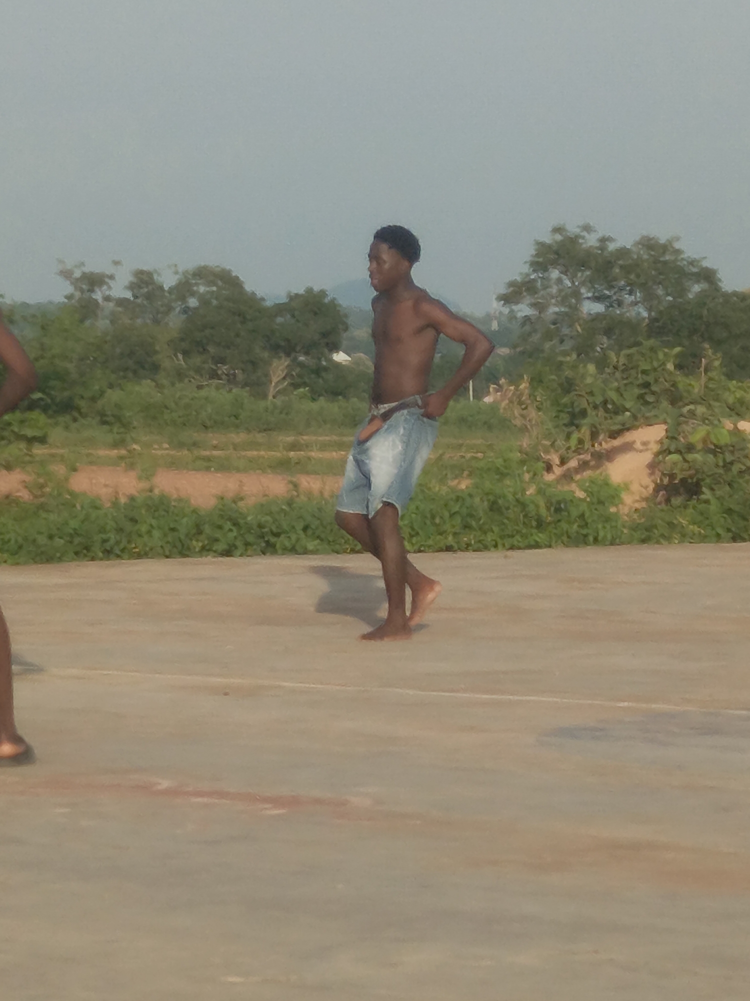 The image depicts an outdoor scene, likely in Akwanga, Nigeria, with a young Black male in the foreground. He is shirtless and wearing denim shorts, with his right hand on his hip and his left arm bent. He is facing towards the left side of the frame. Behind him, there is lush green vegetation and trees, with a hazy, pale blue sky overhead. The ground in the foreground appears to be a paved surface, possibly a court, with some faint markings. The overall impression is of a casual, possibly warm-weather setting.