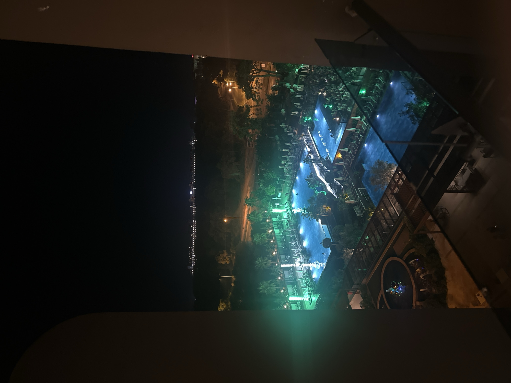 This is a nighttime, high-angle, interior shot from an arched doorway overlooking a resort complex in Manavgat, Turkey. The main focal point is a brightly lit swimming pool area, illuminated by a mixture of blue and green lights. Numerous lounge chairs with umbrellas are neatly arranged around the pool. In the foreground, a circular fountain with colorful lights is visible, along with some surrounding greenery. Beyond the pool, a pathway with streetlights winds through trees, leading towards a distant cityscape with illuminated buildings. The overall impression is one of a vibrant, well-maintained resort at night.