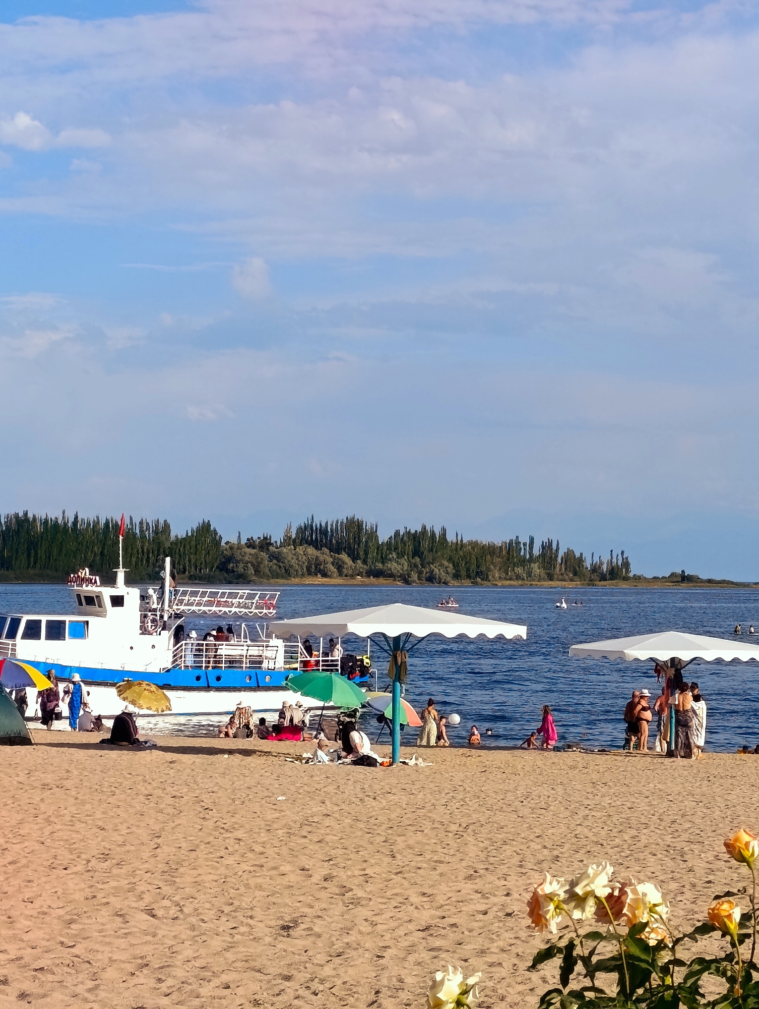 The image depicts a sunny day at a beach near Bayet, Kyrgyzstan. In the foreground, a sandy beach is populated with beachgoers relaxing under umbrellas and tents. Several people are visible, some sitting and others standing. A blue and white passenger boat is anchored in the clear blue water, with people on its deck and swimming nearby. The background features a line of tall, slender trees and distant mountains under a partly cloudy sky. The scene conveys a leisurely atmosphere of a day at the lake.