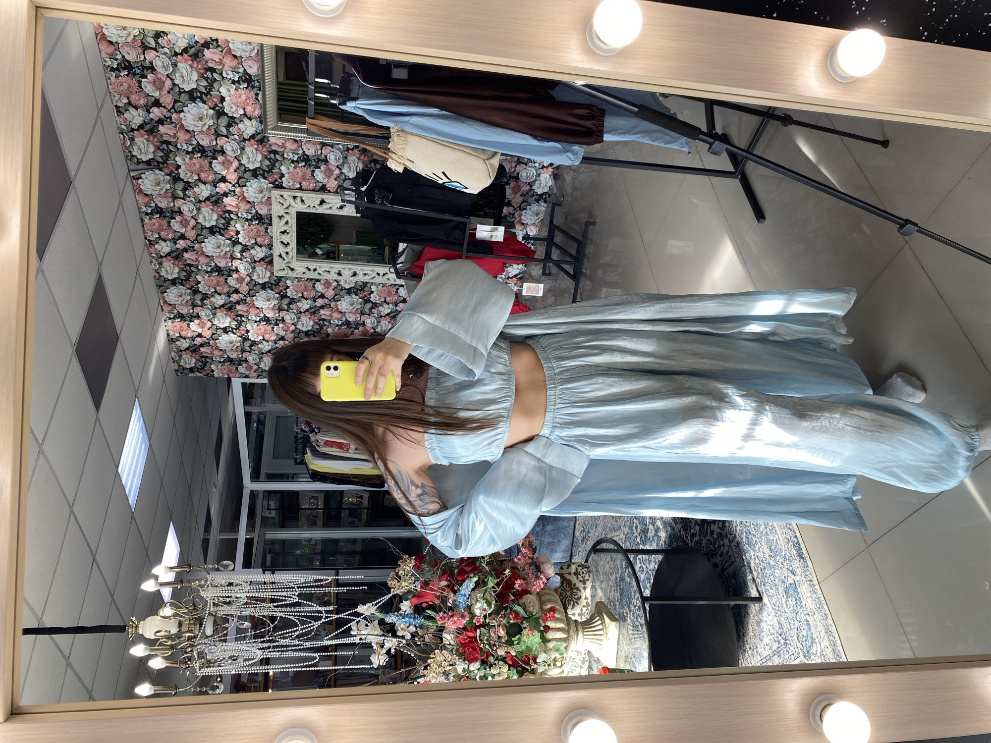 A person is captured in a mirror selfie, standing within what appears to be a clothing store, boutique, or dressing room in Votkinsk, Russia. The individual, seen from the mid-chest down, has long brown hair and is holding a bright yellow smartphone to take the photo. They are wearing a light blue, shiny two-piece outfit consisting of a cropped top with wide, flowy sleeves and a matching long skirt. A prominent tattoo is visible on their left shoulder/upper arm.
The mirror, framed with light-colored wood, features exposed light bulbs along its top and sides, illuminating the scene. Reflected behind the person are various elements of the interior: a wall adorned with dark floral wallpaper featuring pink, white, and green flowers, and an ornate white-framed mirror hanging on it. Clothing racks display garments like brown trousers, a light blue top, and a black top, along with a beige woven bag. A black tripod or stand is also visible on the tiled floor.
To the person's right, a large, colorful artificial floral arrangement in a decorative urn sits on a dark, round table, next to a blue and white patterned rug. Above, a chandelier with multiple lights and strings of pearls or beads hangs, and bright rectangular fluorescent lights are recessed into the ceiling. The floor is light-colored and tiled. The person is wearing white socks. The setting suggests a focus on fashion and display. No discernible text is visible in the image.