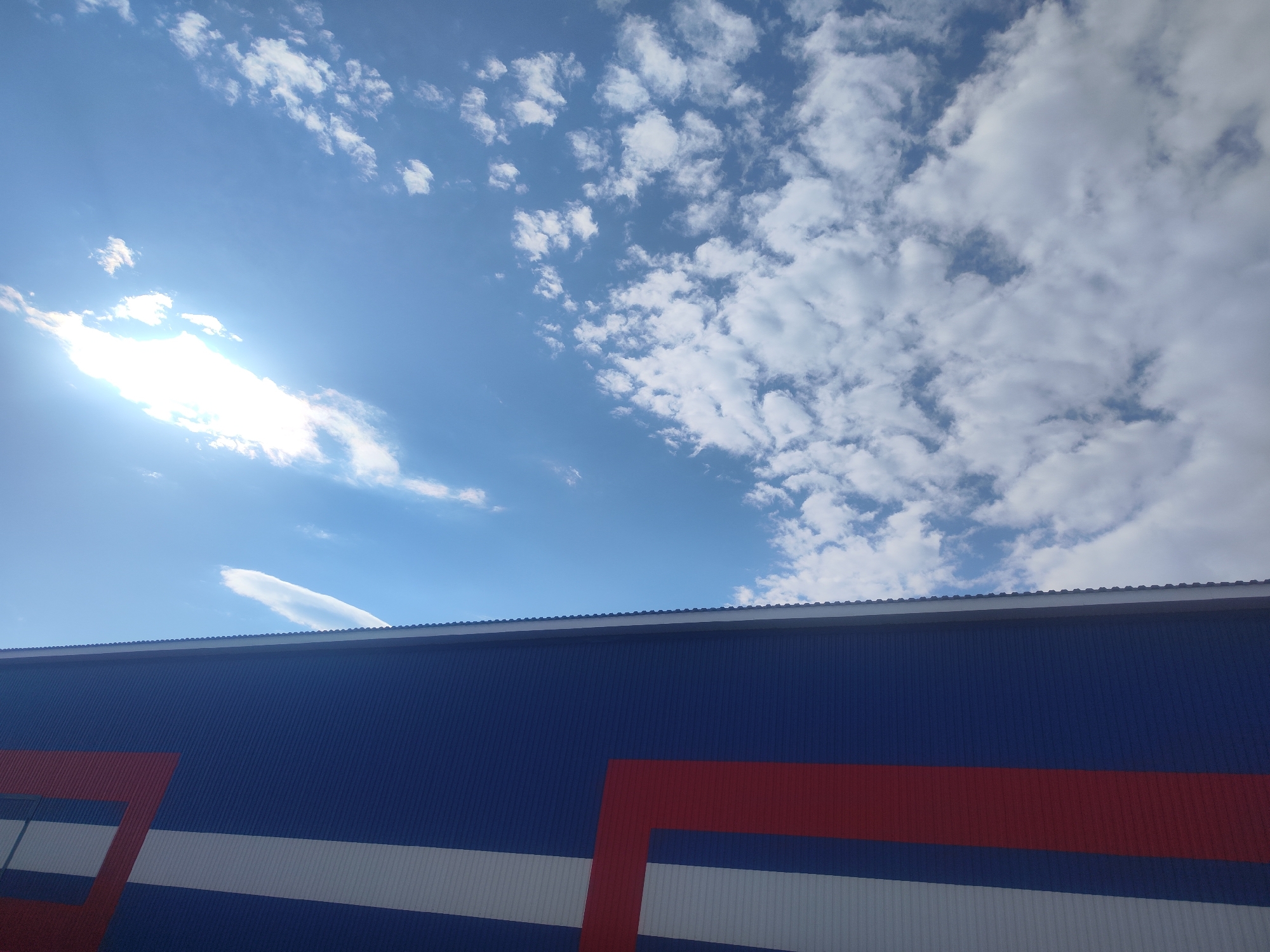 The photograph captures a bright, clear blue sky filled with scattered white cumulus clouds, occupying the upper half of the frame. A strong sun glare is visible in the upper left, emanating from behind a cloud. Below the sky, the upper section of a large, modern industrial or commercial building is depicted. Its facade is constructed from dark blue corrugated metal paneling, with a clean, simple roofline marking the transition to the sky. A distinctive design adorns the building: a broad white horizontal stripe extends across the lower portion of the blue wall, and a striking red geometric element, resembling a partial frame or abstract letter, overlaps both the blue and white sections. A thin, elongated cloud can be seen trailing horizontally just above the building's roofline. The bright illumination indicates a daytime setting with good weather. The scene is located in Chaikovsky, Russia. No people or specific activities are visible, and there is no discernible text.