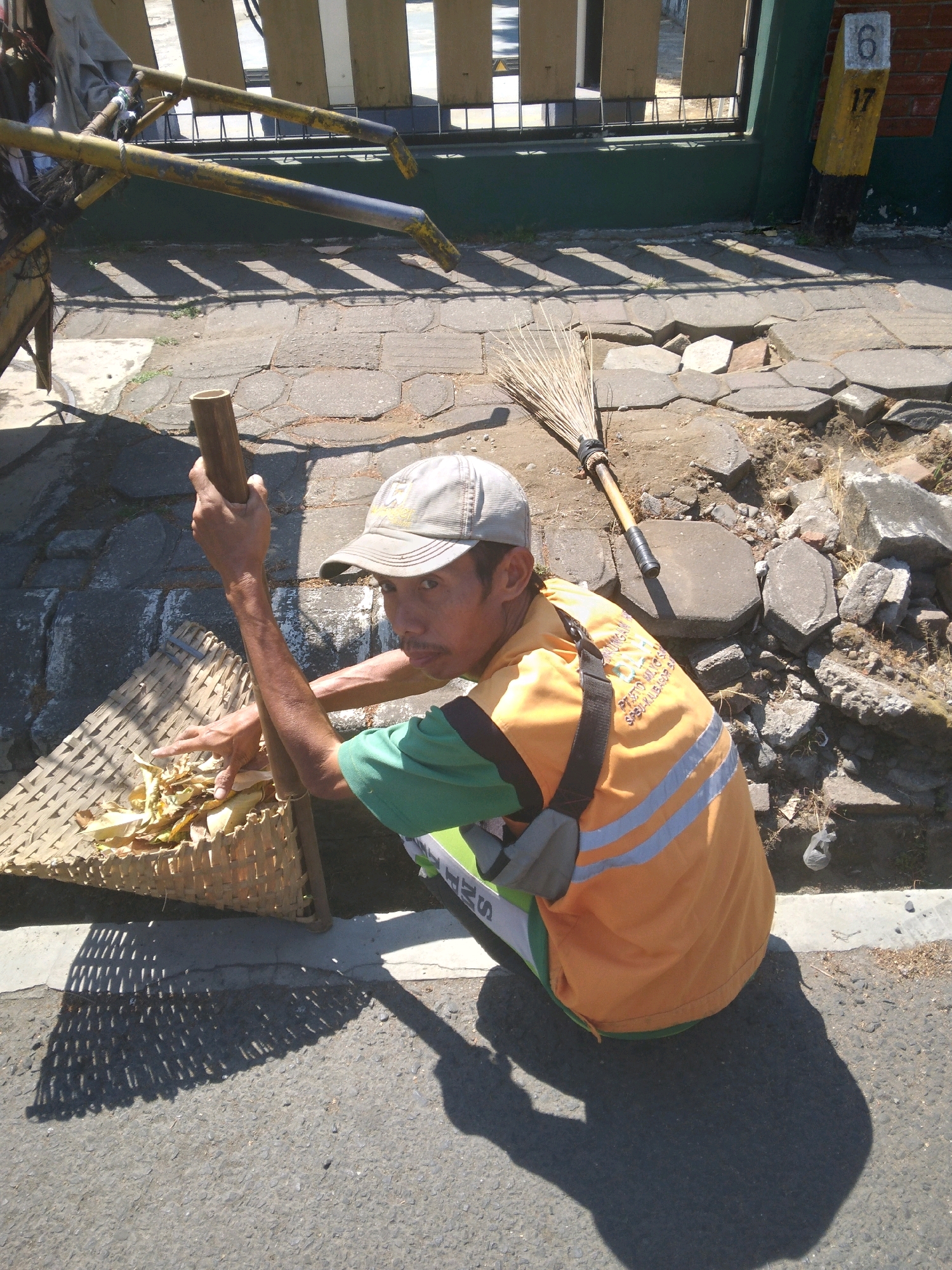 A street cleaner, wearing a light-colored cap, a green t-shirt, and an orange safety vest with reflective stripes, is squatting on a cracked asphalt and paving stone surface. He is holding a wooden pole in his right hand and is using his left hand to point at dry leaves and debris in a woven basket. A broom made of dried grass and a wooden handle lies on the ground nearby. The environment appears to be outdoors, in an urban setting, with a metal fence and a building visible in the background. The scene is illuminated by bright sunlight, casting distinct shadows. The location is identified as Bancarkembar, Indonesia, suggested by the presence of a yellow and black striped pole with the number "6" and "17" on it, a common sight in Indonesian street numbering systems.