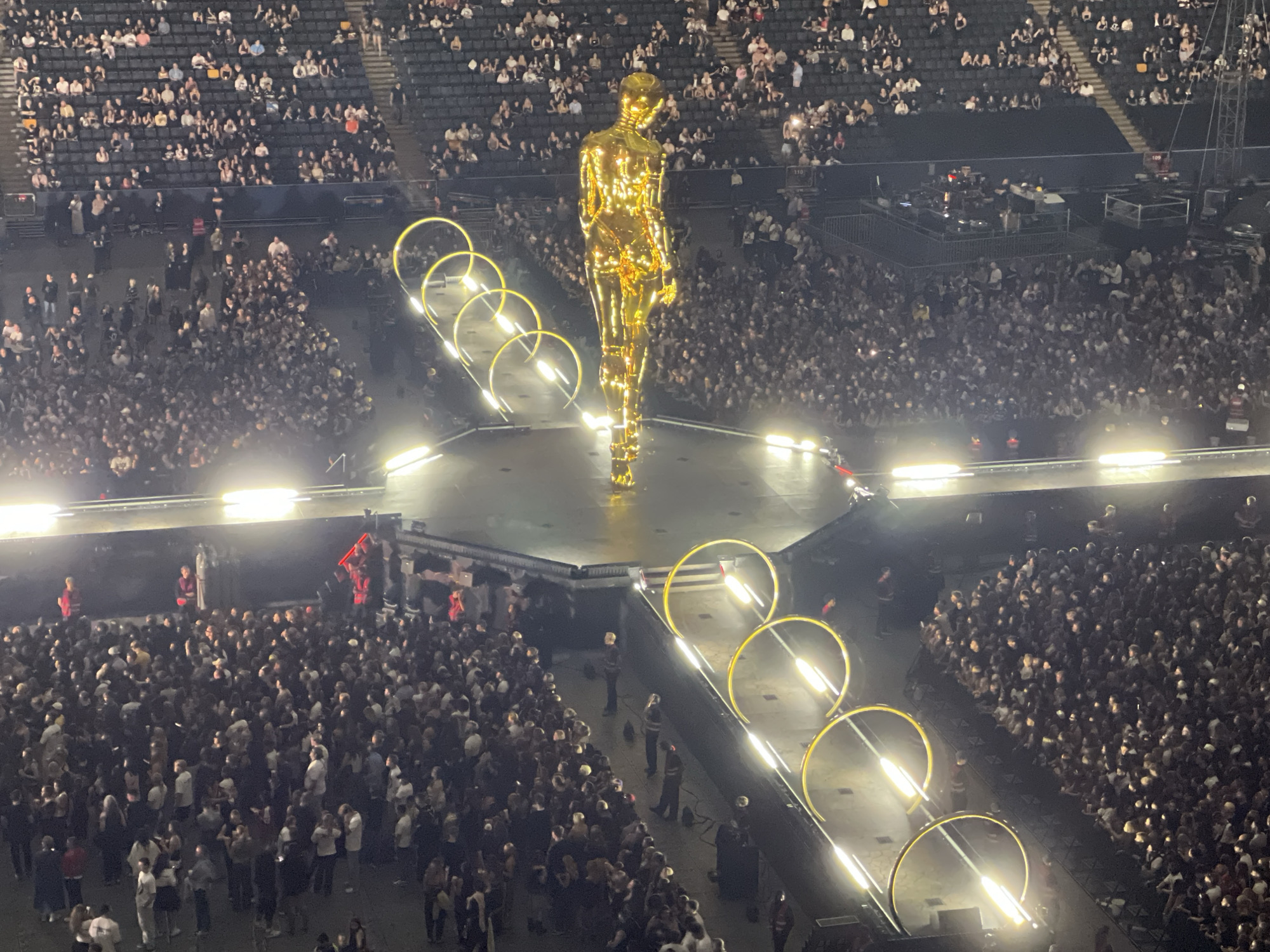 An aerial, high-angle, wide shot captures a large indoor concert or event at the Friends Arena in Solna, Sweden. The central focus is a colossal, golden, humanoid statue standing on a raised stage. The statue is illuminated and reflects the bright lights of the arena. Surrounding the stage are several illuminated, gold, circular structures. A massive crowd of spectators fills the foreground and the tiered seating areas that ascend in the background. Many audience members are visible, some holding up phones to capture the event. The overall atmosphere appears to be one of a large-scale entertainment production.