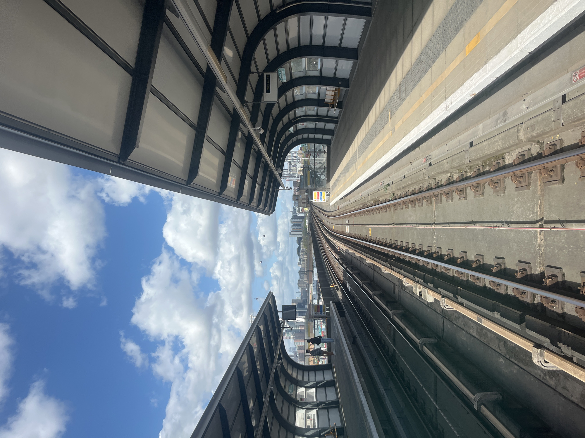 The image captures a view from the Docklands Light Railway (DLR) in London, United Kingdom, specifically looking down the train tracks towards the city skyline. The elevated tracks are flanked by covered station platforms on either side. The platform on the right is visible with its striped yellow and white safety markings. The tracks converge towards a backdrop of buildings, indicating a forward direction of travel into or towards the city center. A blue sky with scattered clouds is visible between the platform coverings.