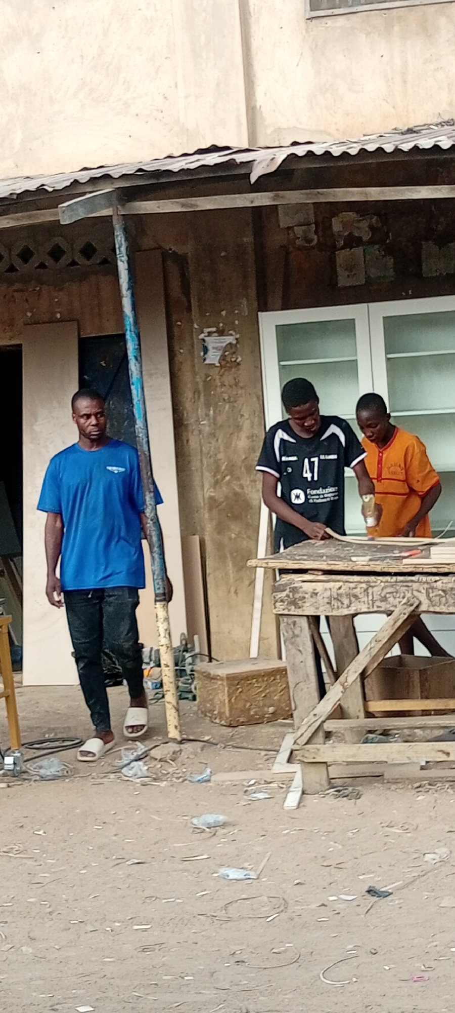 Outside a weathered building in Gama, Nigeria, two young men wearing casual clothes work together at a wooden workbench, appearing to glue pieces of wood. One wears a black and white jersey with the number "47" on it, while the other wears an orange shirt. A man in a blue shirt and black pants, wearing sandals, is walking past them, holding a metal pole. Behind them, against the building, are a wooden panel, white cabinets, and various tools and materials. The ground is dusty and littered with debris.